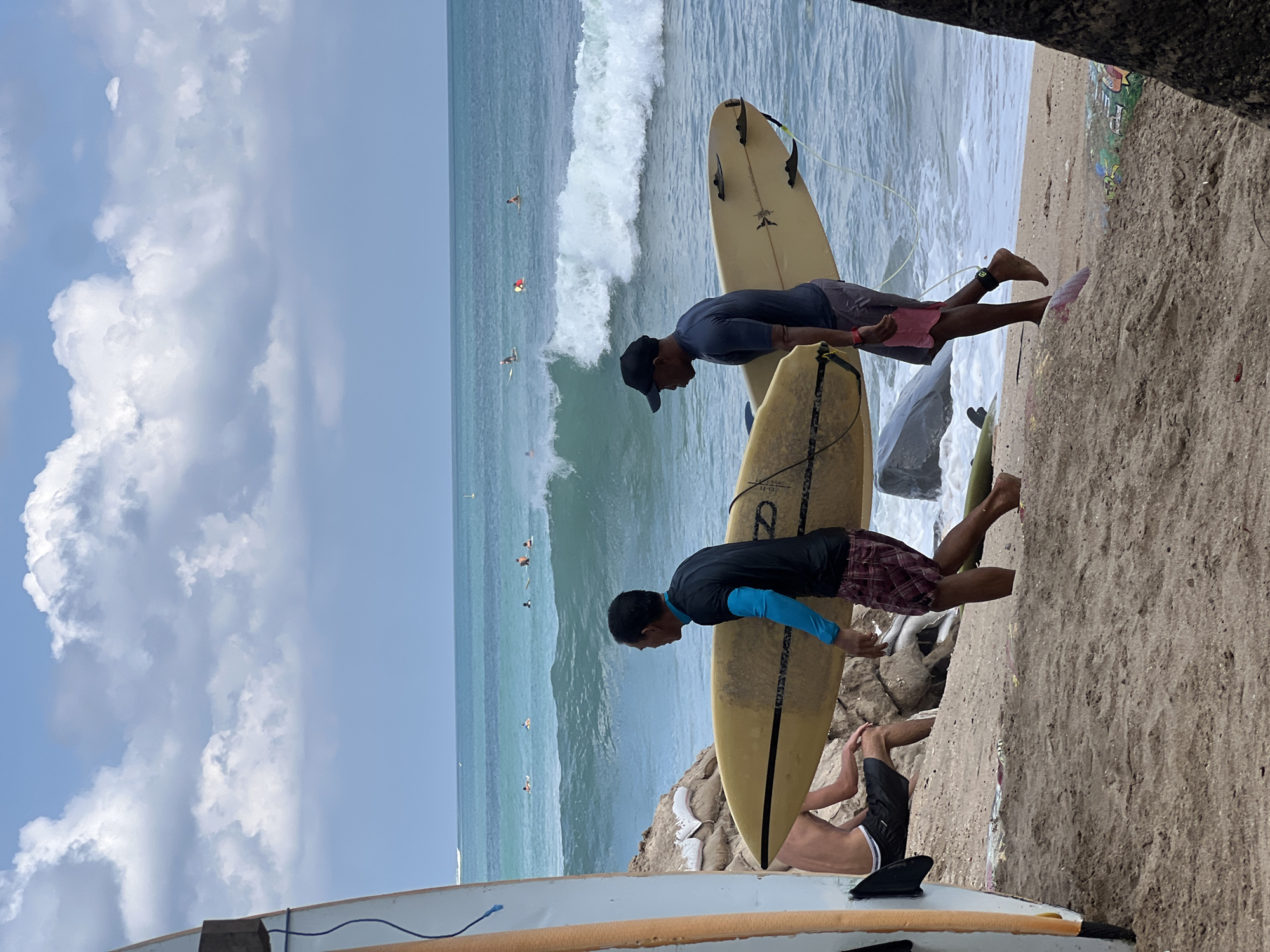 The image captures a sunny day at Kuta Beach, Indonesia. In the foreground, two surfers are walking along the sandy shore, carrying a yellow surfboard between them. The surfer on the left is wearing a blue long-sleeved rashguard and checkered shorts, while the surfer on the right has on a dark t-shirt, shorts, and a black baseball cap. Behind them, the turquoise ocean is active with waves, and several other surfers are visible in the water, some paddling and others riding waves. The sky above is a mix of blue and fluffy white clouds. The scene suggests a typical day at a popular surf spot.