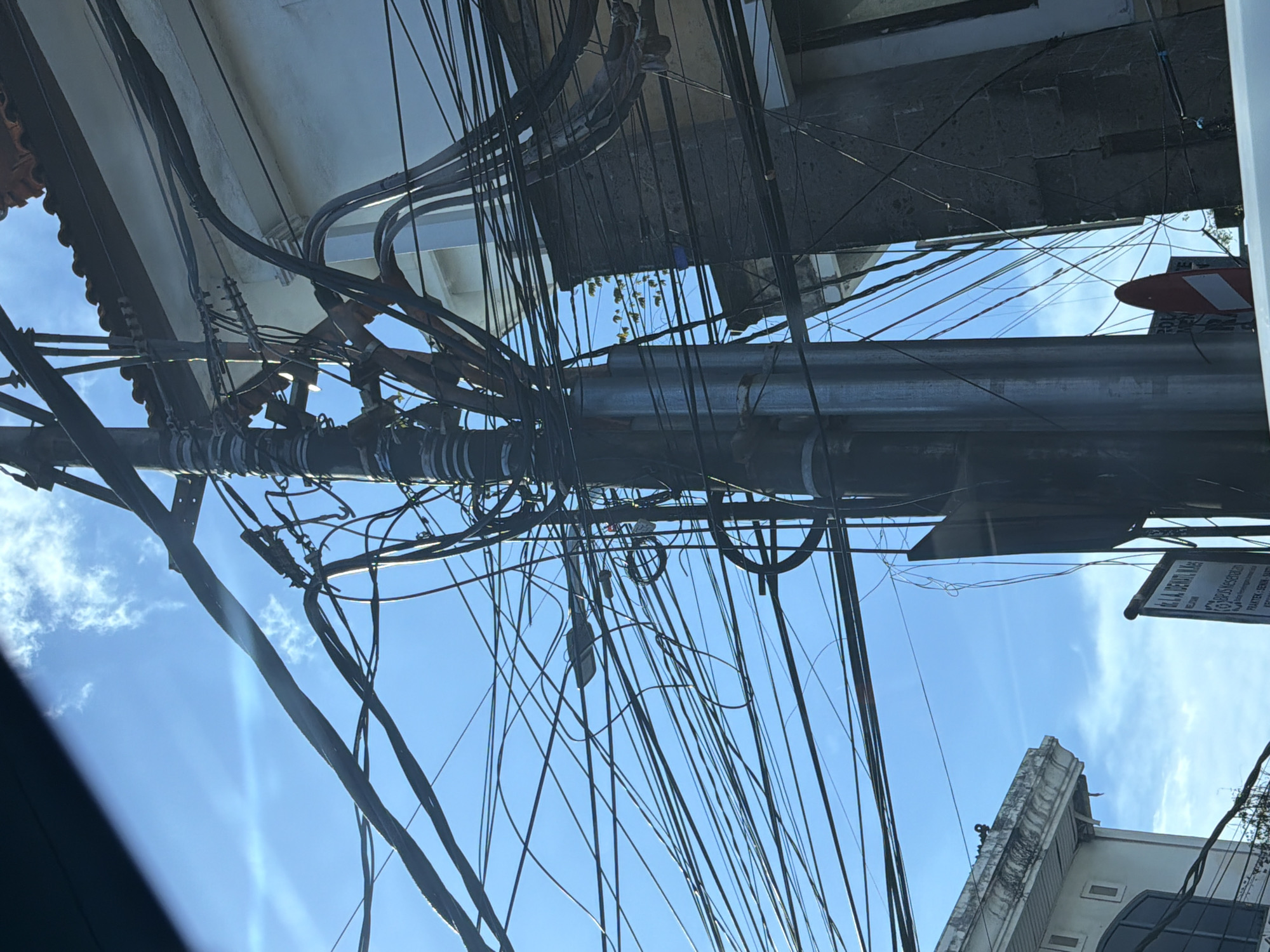 The image captures a dense and chaotic network of overhead utility cables and wires, viewed from below against a bright blue sky with scattered white clouds. The scene is dominated by numerous black electrical and communication lines, which are intricately tangled, looped, and draped across the frame.
A large, grey utility pole or structural column runs vertically through the center-left, acting as the primary support for many of these wires. It is equipped with multiple clamps, attachments, and conduits holding the various bundles of cables. The sheer volume and disorganization of the wires suggest an urban or semi-urban environment.
In the background, fragments of buildings are visible. These include light-colored concrete structures, one with a distinctive upper section featuring architectural details like a cornice or roofline. A dark window is also partially seen. The bright conditions indicate a sunny daytime.
On the upper right, a vertical, dark rectangular sign is prominent. Readable text on the sign identifies it as "JL. RA. N. DEWI" (likely a street name, common in Indonesia), followed by a phone number "081915998270". Further text includes "Klinik," "Praktek," and "Dokter Gigi" (Clinic, Practice, and Dentist), along with "Spesialis Orthodontic" (Orthodontic Specialist), clearly indicating a dental clinic. Below this sign, a red and white striped barrier or sign is partially visible.
No specific activity or individuals are depicted; the image is a static representation of a common infrastructure sight in many parts of Southeast Asia, consistent with the given location context of North Kuta, Indonesia.