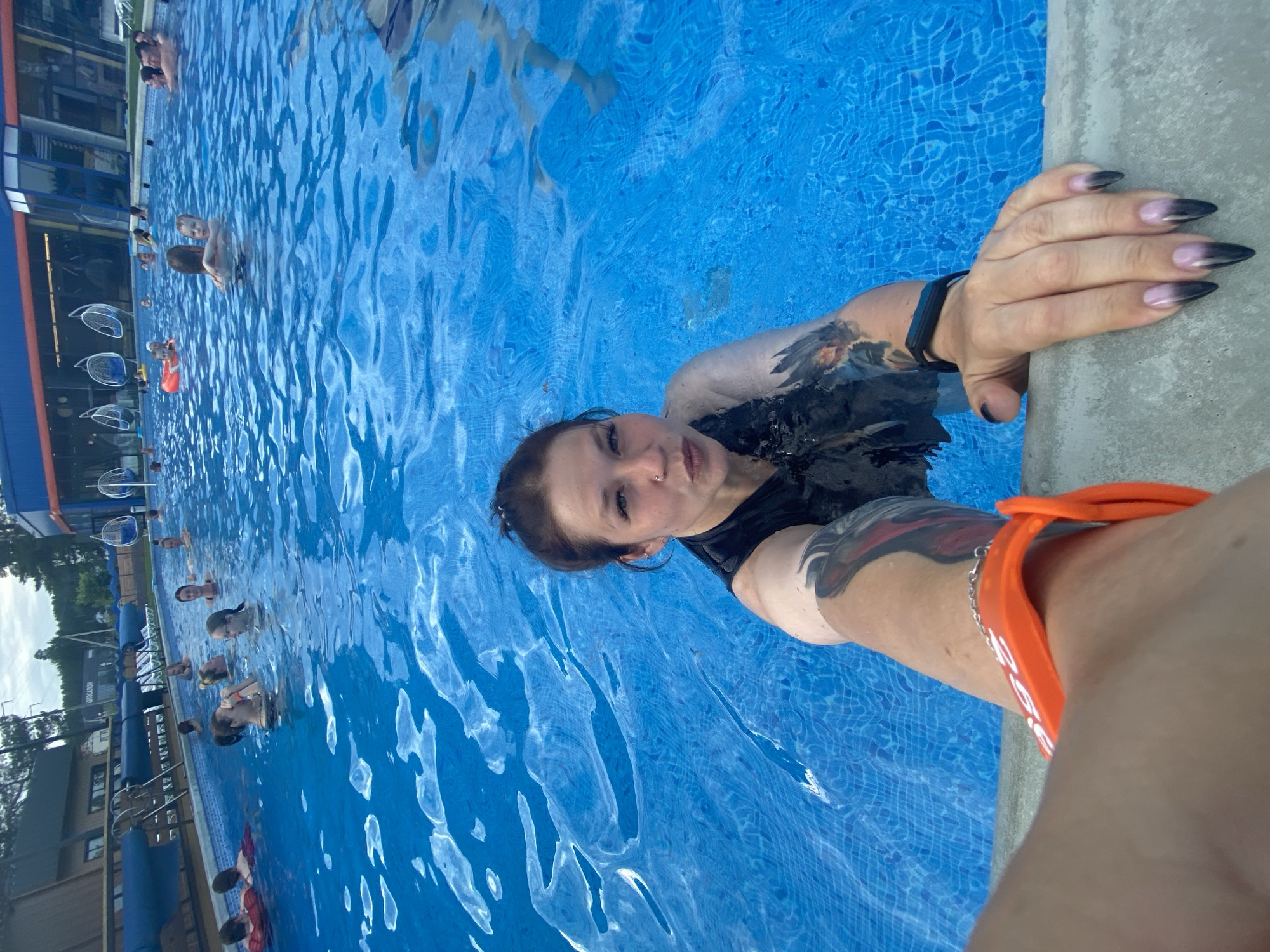 This is a selfie taken in a crowded outdoor swimming pool. The perspective is from the water's surface, looking up towards the person taking the photo and others in the pool. The water is a vibrant blue, with sunlight reflecting off its surface, creating rippling patterns.
The main subject is a young woman with dark hair and visible tattoos on her arm and shoulder, wearing a dark swimsuit. She is holding onto the edge of the pool, which appears to be made of concrete. Her hand, with pointed black and pink gradient nails, is resting on the pool's edge. She is looking directly at the camera with a playful or engaging expression. She is also wearing an orange wristband with white lettering.
In the background, several other people are visible in the pool, enjoying themselves. Some are swimming or wading, while others appear to be relaxing. The pool area has a modern aesthetic with blue and orange accents visible in the building structure behind the pool. There are also some hanging chairs or loungers by the side of the pool.
The overall mood seems to be relaxed and fun, indicative of a summer day spent at a water park or public pool. The lighting suggests it is daytime, likely sunny. While the location is identified as Votkinsk, Russia, there are no specific visible cues in the image that confirm this without prior knowledge. The water is clear and inviting, and the sun's reflection indicates good weather. The orange wristband on the woman's arm may be an identifier for entry or a locker, common in such recreational facilities.