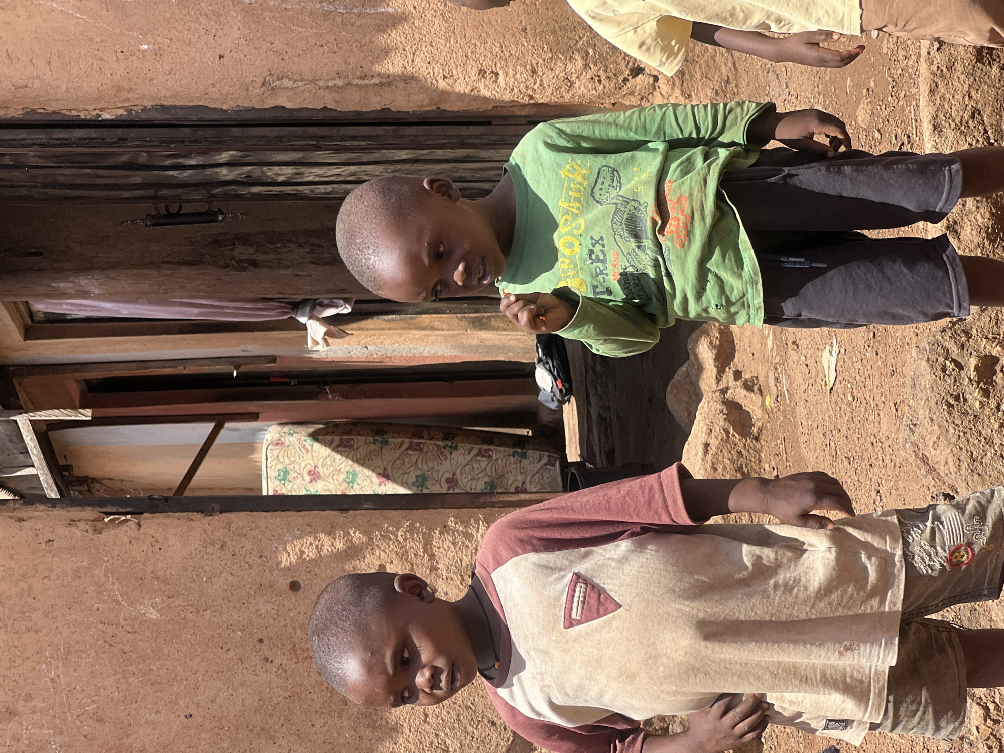 Two young boys stand outside a modest dwelling in Gura Top, Nigeria. The older boy is wearing a light-colored shirt with maroon sleeves and a small pocket, and patterned shorts. The younger boy has on a green shirt with a dinosaur print and dark-colored shorts. The building behind them appears to be made of mud or clay, and there is a mattress visible through a window or opening. The ground is dry and dusty.