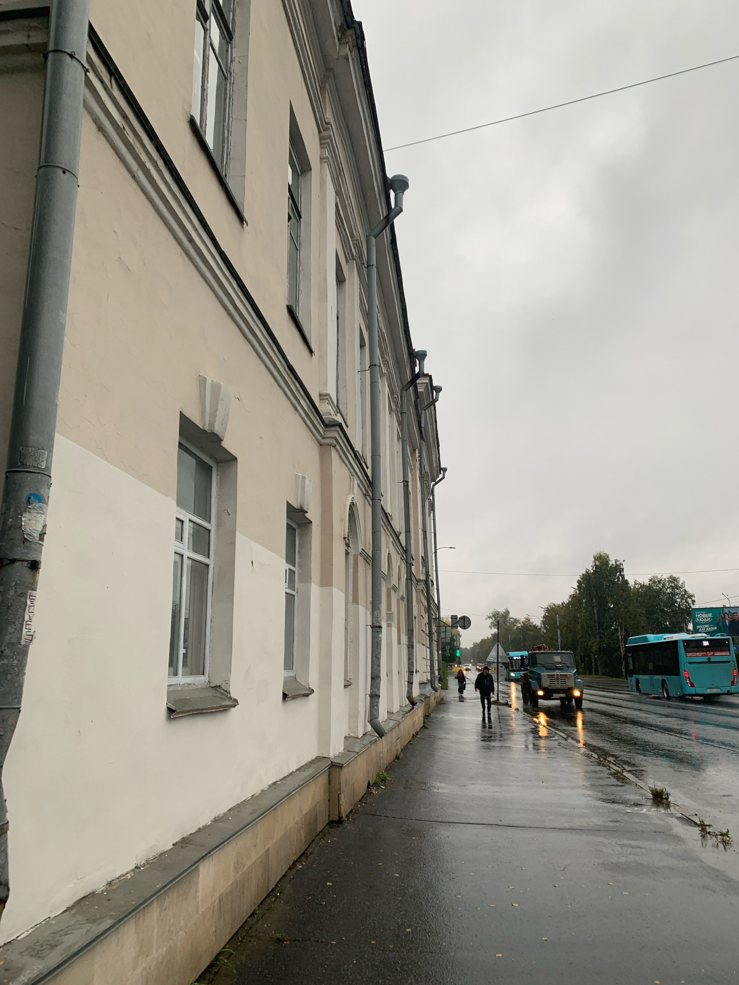 This is a horizontal, eye-level, medium shot of a street scene in Arkhangelsk, Russia, on an overcast day with rain. The left side of the frame is dominated by the light-colored facade of a multi-story building, featuring a prominent vertical pipe and regularly spaced windows. The building's lower half is a lighter shade than the upper half. A wet sidewalk runs along the building, reflecting the dull sky and the lights of passing vehicles.
In the background, a street is visible with traffic. Several vehicles, including buses and a truck, are moving along the wet asphalt. The road surface is dark and reflective due to the rain. Two pedestrians are walking on the sidewalk in the mid-ground. One person, wearing a dark jacket and pants, is walking towards the camera, while another person, in dark clothing, is walking away from the camera. The overall atmosphere is somber, likely due to the weather. A billboard is visible on the right side of the background, featuring text and images.