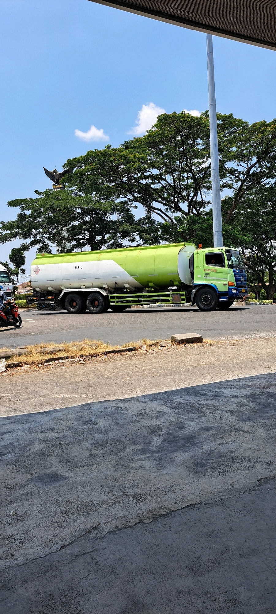 A bright green and white tanker truck is parked on the side of a road in what appears to be Pandaan, Indonesia. The truck is a Hino, indicated by a logo on the side of the cabin. The truck's trailer is a large cylindrical tank, painted white with a green stripe running along the top. On the side of the trailer, the letters "P.S.C." are visible in black. The truck is parked on an asphalt road, with a dry, grassy shoulder. In the background, there are lush green trees and a bright blue sky with a few white clouds. A tall, metallic pole is also visible in the background, possibly a flagpole or utility pole. To the left of the tanker truck, a motorcycle with a rider is partially visible, indicating the presence of other vehicles and activity. In the far background, a large sculpture of an eagle with outstretched wings is mounted on a pedestal. The overall scene suggests a sunny day in a semi-urban or roadside environment.