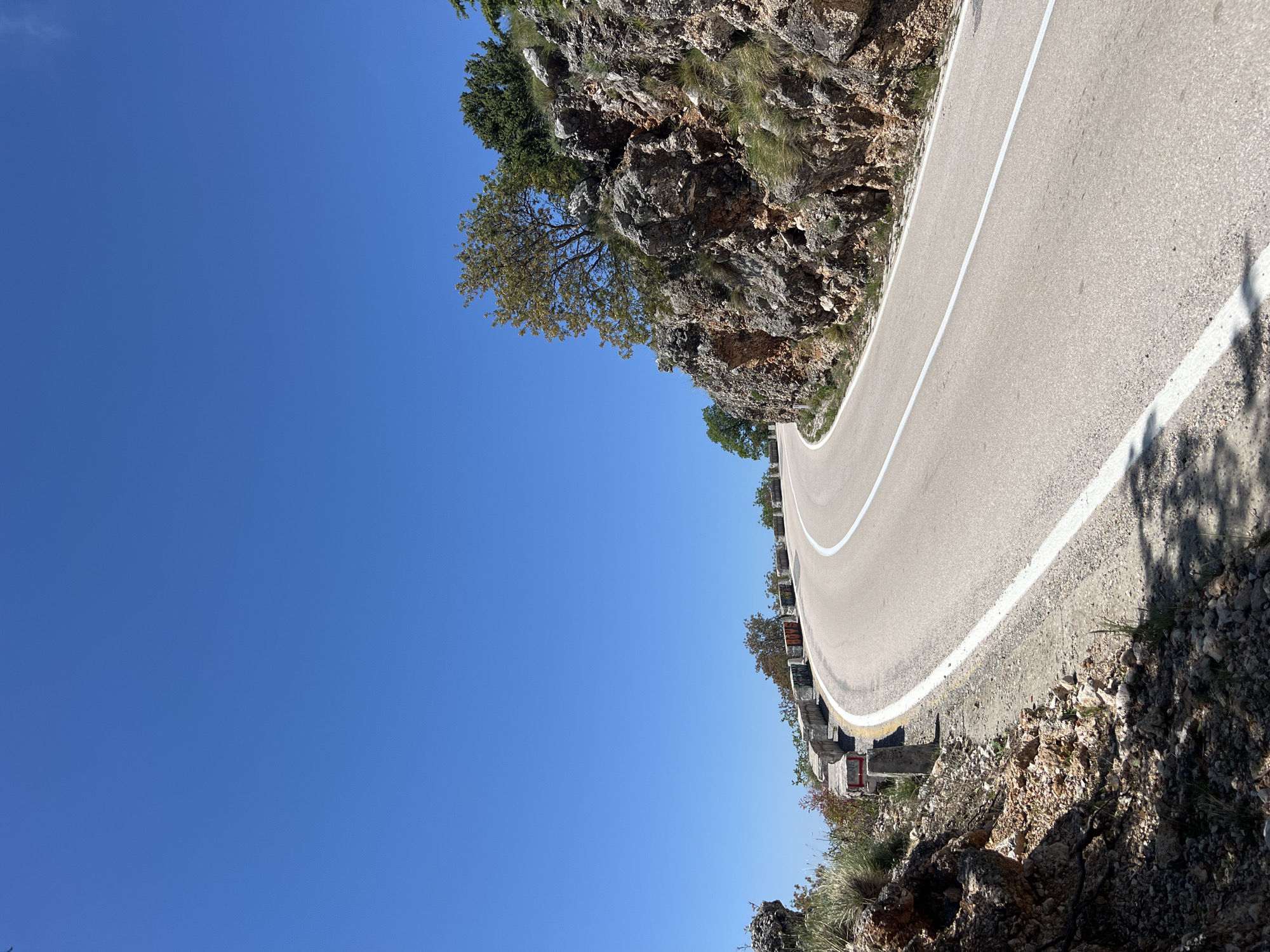 The image displays a scenic view of a winding asphalt road cutting through a mountainous landscape under a clear, vibrant blue sky. The road features a solid white line marking its outer edge and a dashed white line down its center, curving gently to the right and receding into the distance.
To the left of the road, a steep, rugged rock face rises, composed of large grey and brown boulders. Sparse green grass, small shrubs, and several trees with green and hints of autumnal foliage cling to the rocky slopes, casting distinct shadows in the bright sunlight.
On the right side of the road, a series of concrete barriers or guardrails indicate a drop-off or steep descent. These barriers are formed from square concrete blocks, some equipped with metal posts and small red or orange reflective markers. Further down and to the right, more rocky terrain and sparse vegetation are visible below the road level.
The scene is captured on a bright, sunny day, likely during midday or early afternoon, characterized by the intense sunlight and a vast, cloudless sky that dominates the upper left portion of the frame. No people or visible text are present in the image. The setting suggests a serene, elevated route, typical of mountain passes or coastal roads in areas like Novoselje, Montenegro.