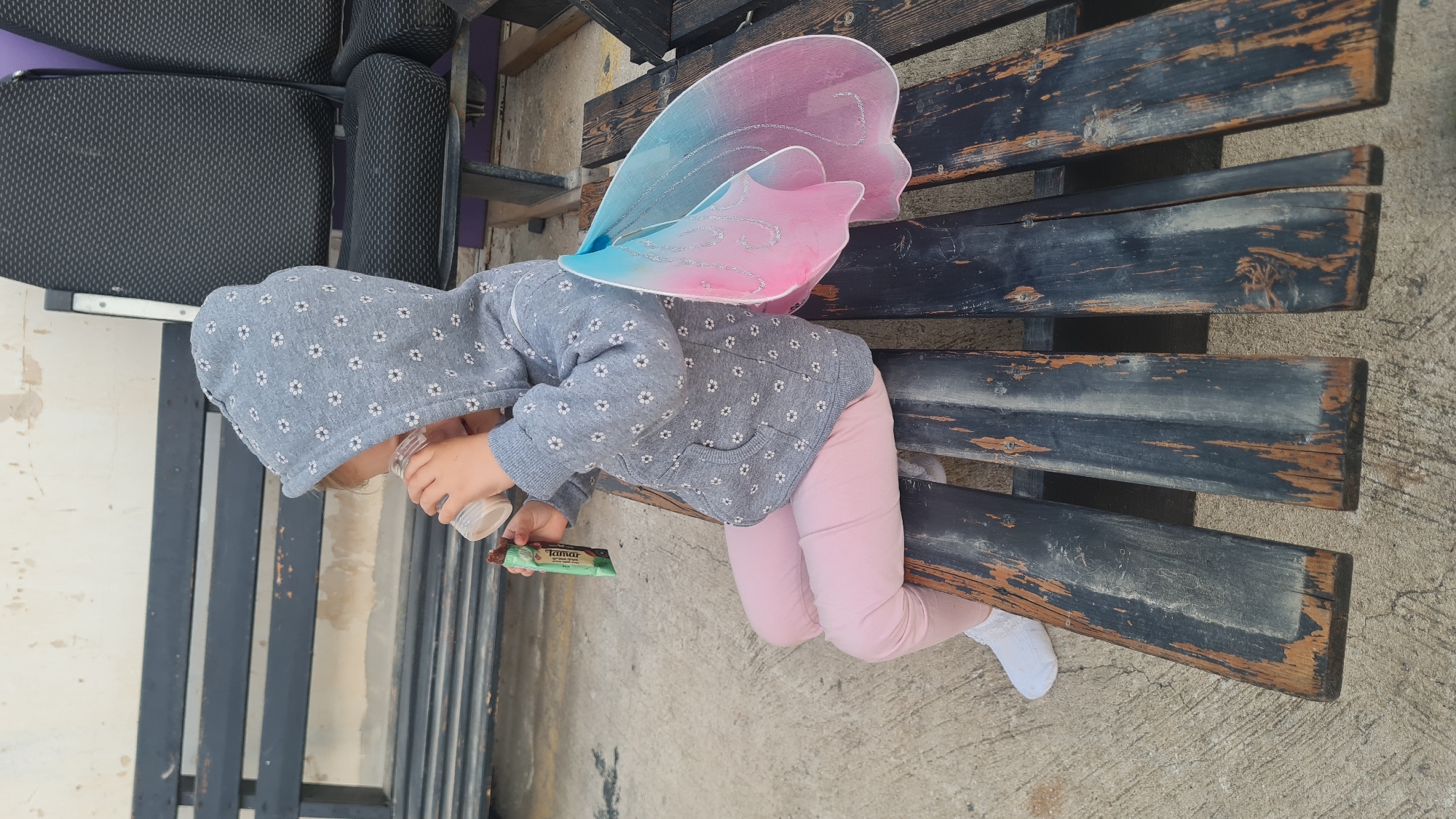 A child wearing a gray hooded sweatshirt with a floral pattern, pink leggings, and white socks is lying prone on a dark, slatted wooden bench. The child has pink and blue fairy wings attached to their back and is holding a small, clear jar in one hand and a green-wrapped snack bar labeled "Tamar" in the other. The bench is located outdoors on a concrete surface. A black chair with a purple seat is partially visible next to the bench. The scene is in Rishon LeZion, Israel.