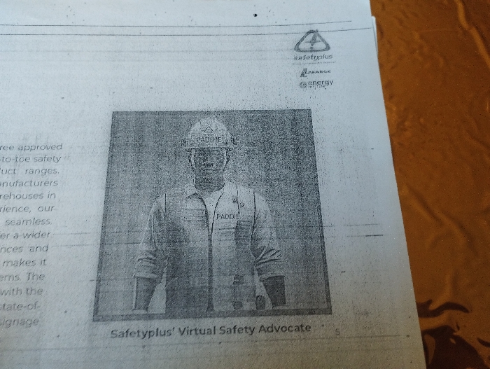 The image shows a grayscale photograph of a man wearing a hard hat and safety vest. The man is standing with his hands crossed. He is looking straight ahead. The image is a part of a printed document. The document has the text "Safetyplus' Virtual Safety Advocate" at the bottom of the image. The coordinates 10.917, 11.476 locate the image in Ashaka, Nigeria. There is no other relevant detail to be observed in the image.