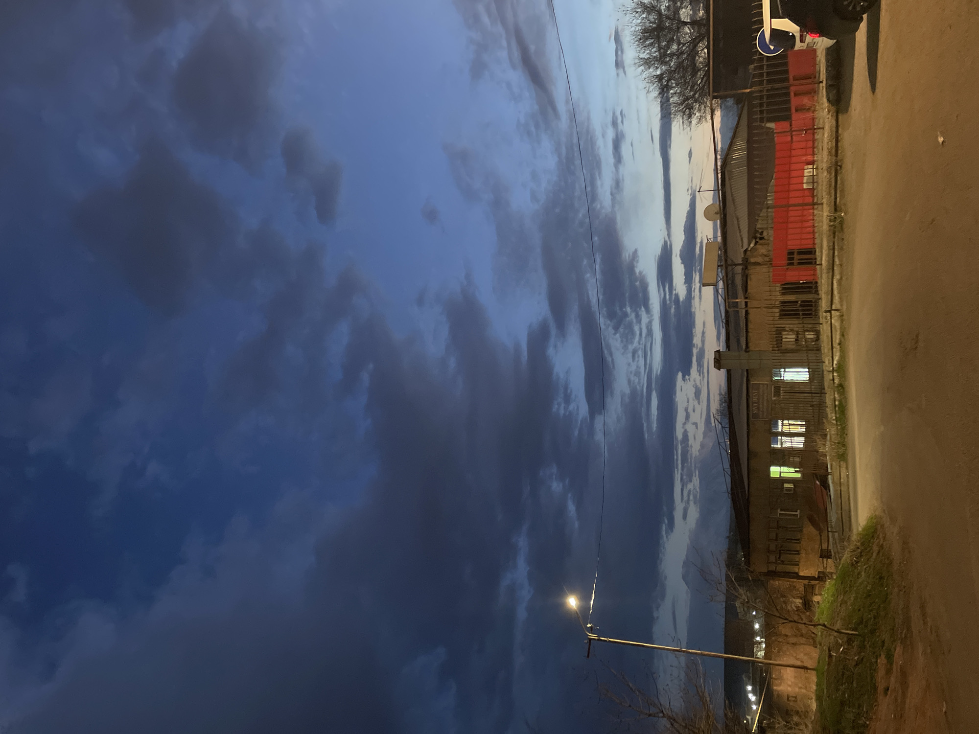 Here is a forensic description of the image:
The image captures a street scene in Yerevan, Armenia, at dusk. The sky is predominantly filled with dark, heavy clouds against a backdrop of fading blue light. On the right side of the frame, a street bordered by buildings is visible. One building appears to have a red exterior with a corrugated metal roof. Another building has illuminated windows, suggesting that it is inhabited or in use. A street lamp is visible, emitting a warm yellow light. The street surface appears unpaved or partially paved with dirt or gravel. The overall scene conveys a sense of urban life transitioning into evening.