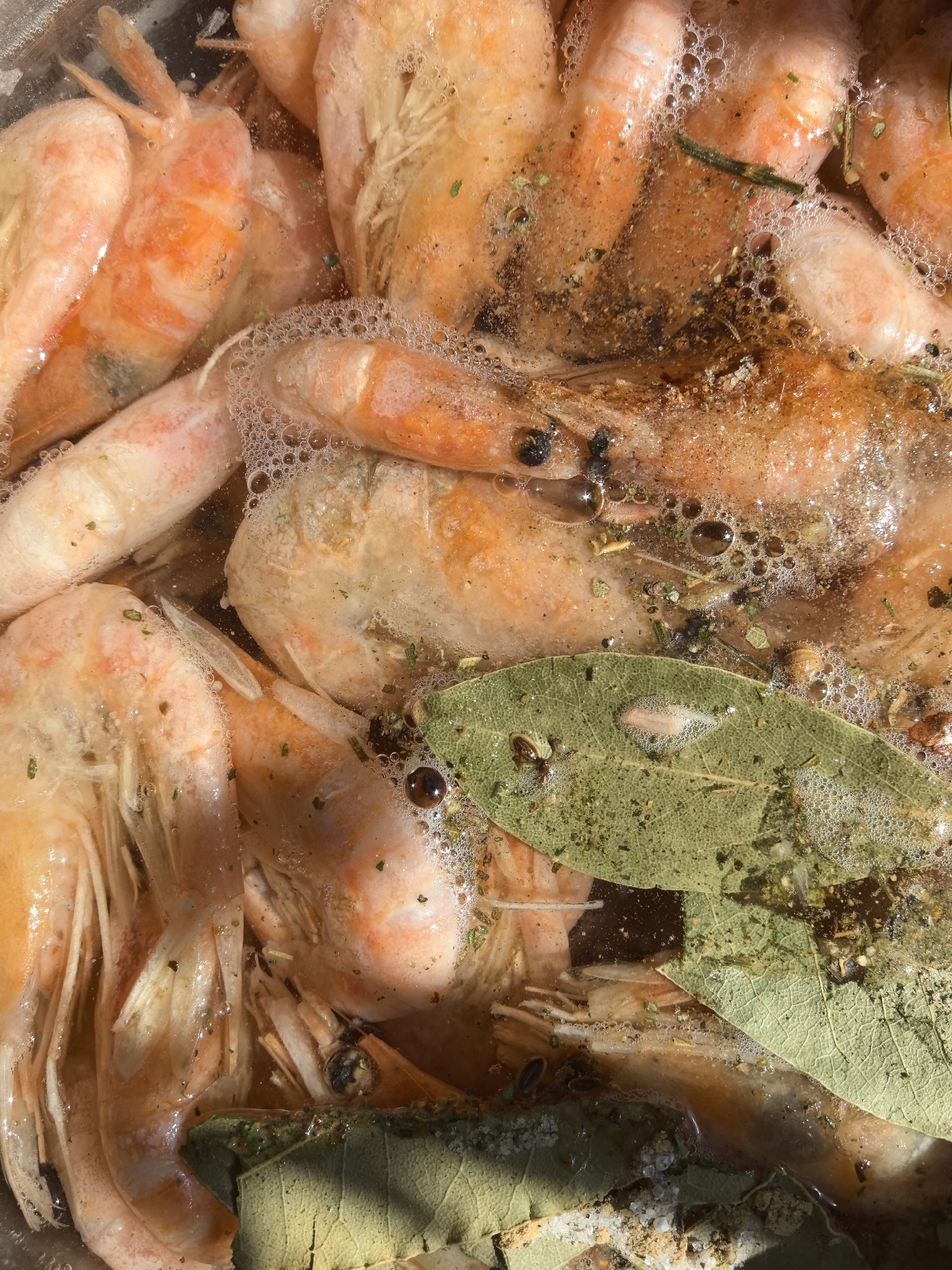 The image shows a close-up view of cooked shrimp, seemingly poached or boiled, with visible bubbles on the surface of the liquid. Bay leaves and various seasonings are mixed in with the shrimp. The shrimp are mostly orange and pink. The composition suggests they are in a container or dish. There is no indication of location or city.
