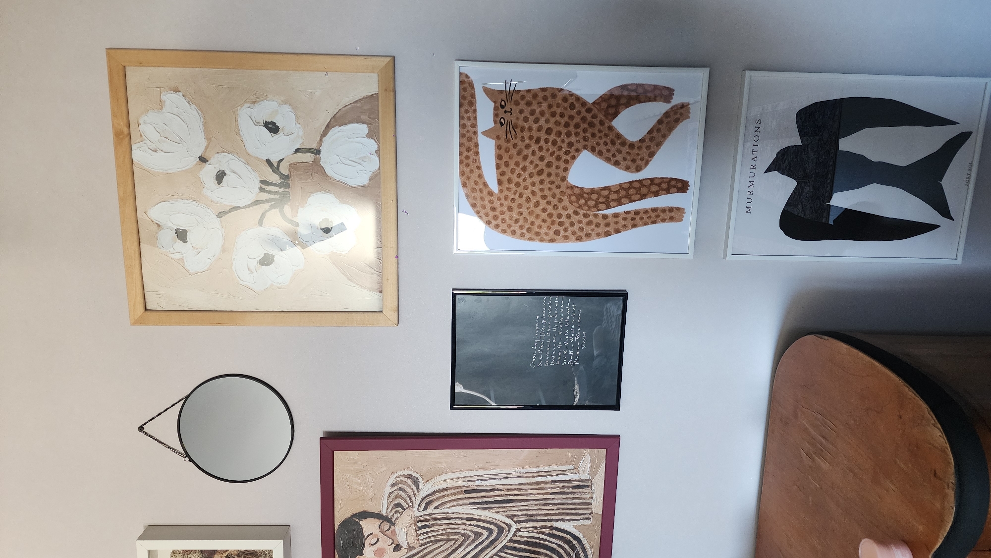 The image shows a wall decorated with various pieces of art and a round mirror. The art includes a floral painting in a wooden frame, a print of a spotted cat in a white frame, a print of a bird with the words "MURMURATIONS" and "SORT SOL" in a white frame, a framed print with text, and a painting of a person in a striped outfit in a maroon frame. The mirror is round with a black frame and hangs from a chain. A wooden shelf is partially visible on the right side of the image. The setting does not appear to be in Smolensk, Russia.