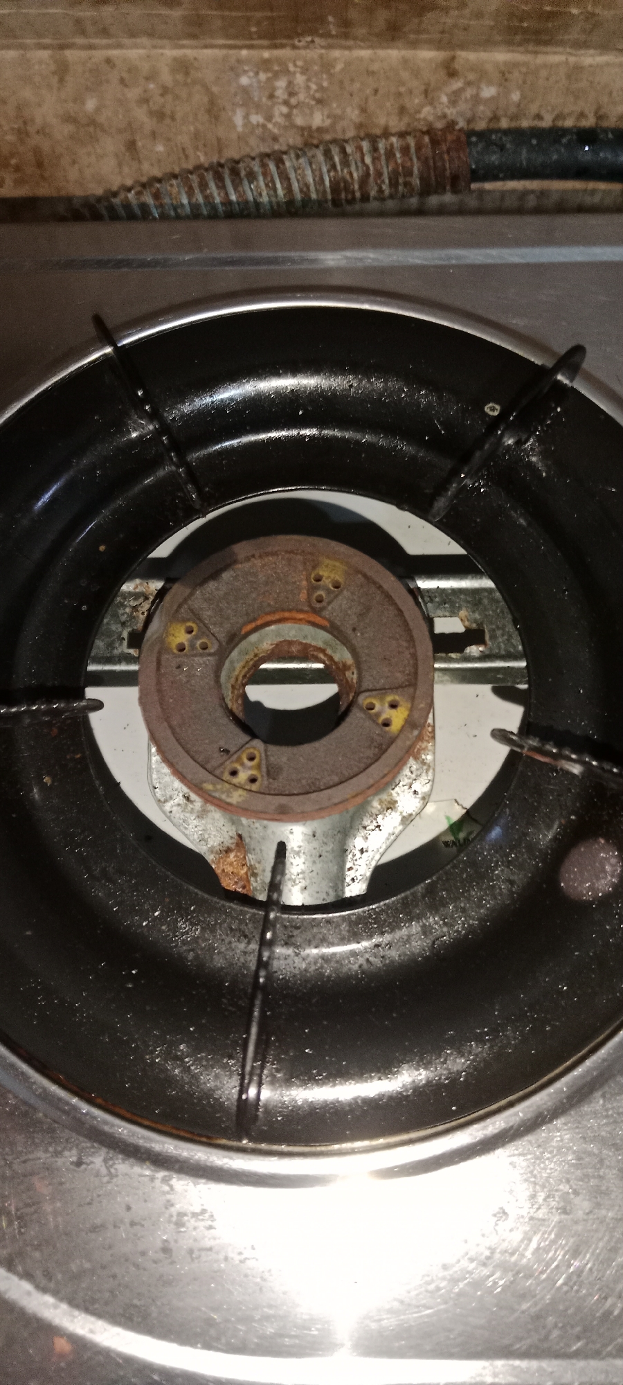 The image shows a close-up, overhead view of a gas stove burner. The burner assembly, made of a rusty, circular metal plate with yellow accents, is situated within a black metal grate. The grate itself is part of a larger stainless steel stovetop. The background consists of a textured, beige surface and a corrugated metal pipe, possibly a gas line, that appears rusted. The overall scene suggests a domestic kitchen environment, likely in Subang, Indonesia, given the context provided. There are no people or discernible actions depicted in the image. The lighting is somewhat harsh, highlighting the texture and wear on the stove components.
