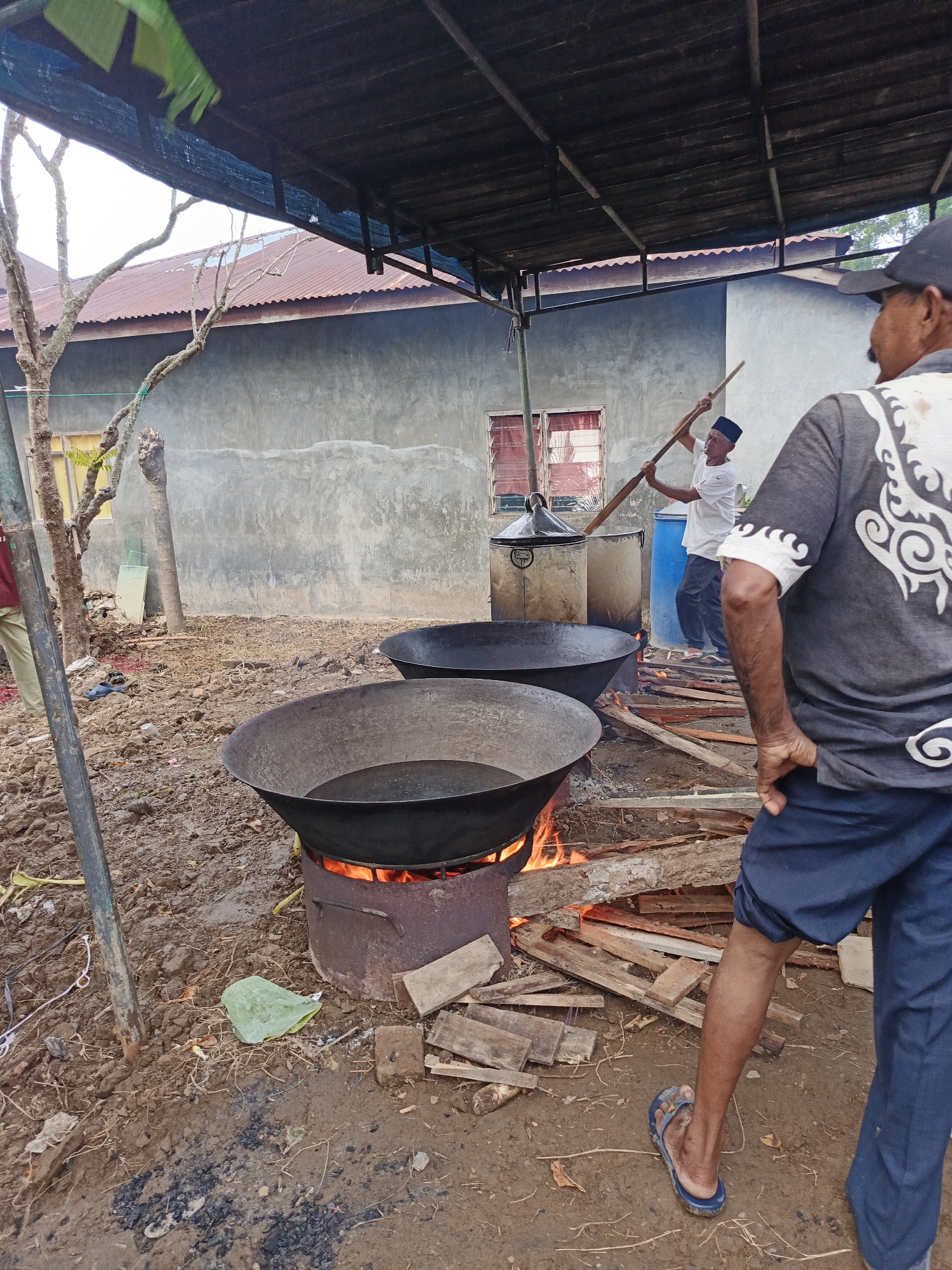 This detailed image captures an outdoor cooking scene in Namplohbaro, Indonesia, under a makeshift shelter. In the foreground, a man is partially visible from the back, wearing a dark grey t-shirt with a white ornate design, dark blue shorts, and blue flip-flops on muddy ground. His right arm is bent, with his hand near his hip.
Directly in front of him, two large, dark-colored woks or cauldrons are positioned on the unpaved, uneven ground. The lower wok is actively heated by an open wood fire contained within a metal drum acting as a stove. A stack of wooden planks and smaller pieces of wood, serving as fuel, lies next to this setup. The wok above it appears to be empty.
In the mid-ground to the left, another man, lighter-skinned, is actively stirring the contents of a taller, large cylindrical pot or container. He wears a white t-shirt, dark pants, and a black cap, holding a long wooden paddle with both hands. A blue plastic barrel is visible behind him. Smoke is faintly discernible rising from the area of the active cooking.
The background features a concrete-walled building with a reddish corrugated metal roof, showing a section of a window with red louvers. Bare tree branches are visible on the far left, extending towards the bright sky. The ground throughout the scene is earthy, muddy in places, with scattered dry vegetation, debris, and some ash. A single green leaf rests on the ground near the lower wok. A metal pole, part of the shelter's structure, stands vertically on the left side of the image.