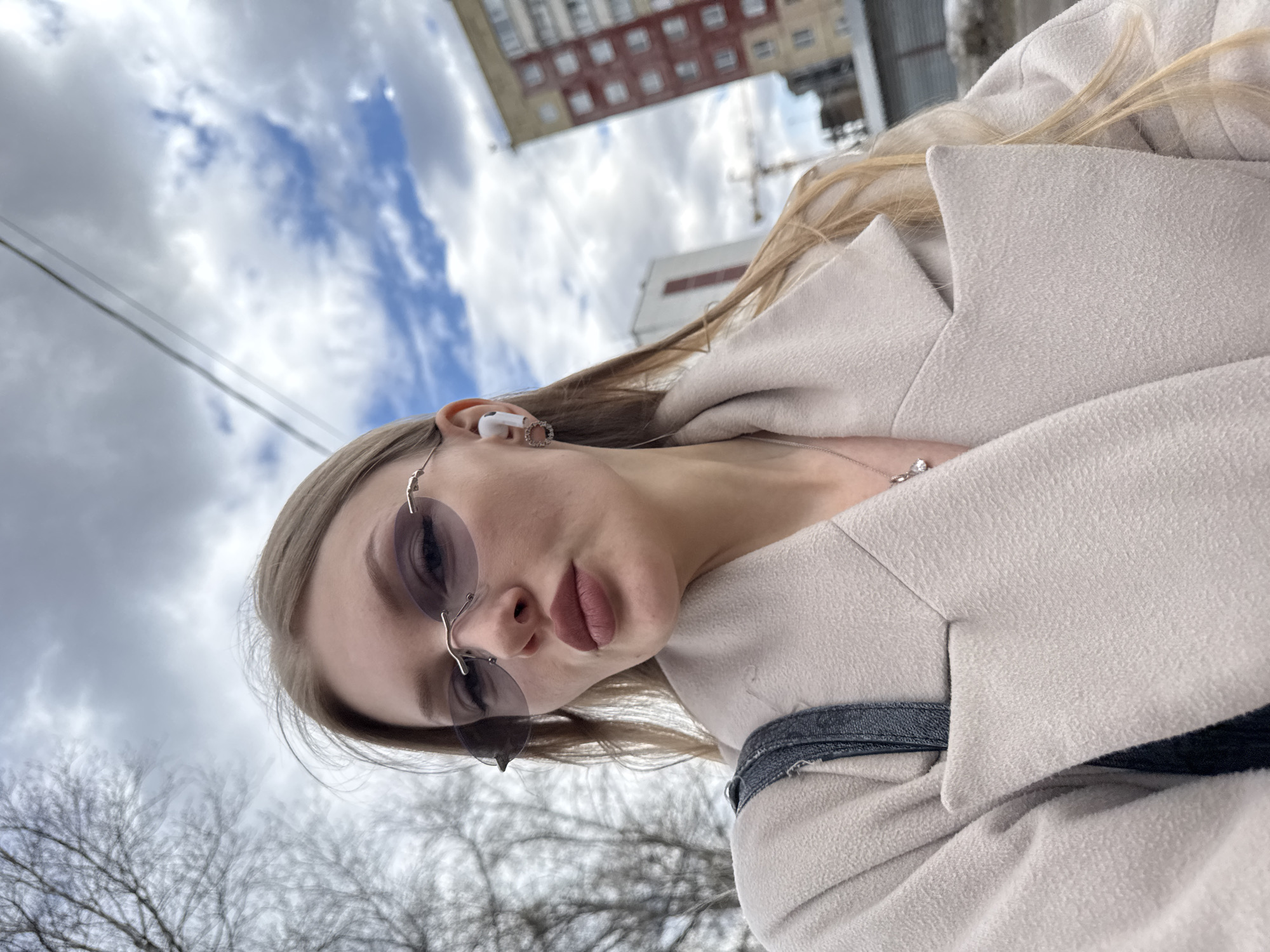 The image is a close-up, low-angle portrait of a young woman, rotated 90 degrees counter-clockwise. She is positioned centrally, looking directly at the camera, with her head tilted to the side. She has long, light blonde hair and is wearing rimless sunglasses with light purple-tinted lenses. A white earbud is visible in her right ear, and a delicate silver necklace with a small pendant adorns her neck. Her full lips are colored with a nude-pink lipstick. She is dressed in a light beige or cream-colored coat, with the collar and shoulder visible.
The background, seen from a low perspective, features a cloudy sky with glimpses of blue. In the upper part of the image (which would be above the woman if the photo were correctly oriented), a multi-story residential building with reddish-brown facades and white windows appears inverted. Further back, another building is partially visible. Bare tree branches are prominent in the lower left, indicating a late autumn or winter season. Overhead utility wires also cross the sky. The scene depicts an urban environment during daytime, under partly cloudy skies, set in Perm, Russia. No visible text is present in the image.