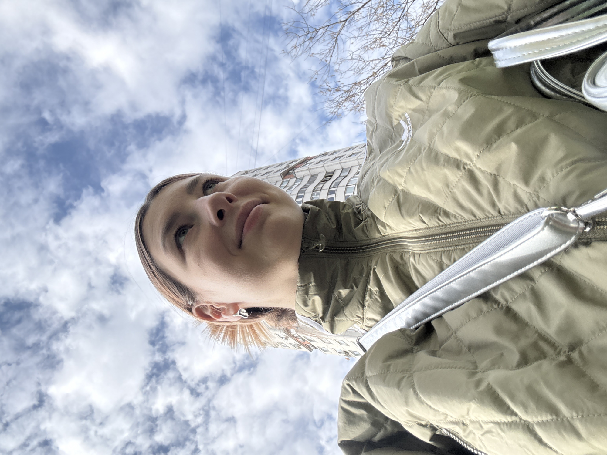 The image shows a low-angle view of a woman wearing a light green quilted jacket with a white strap slung across her shoulder. She is looking upwards and slightly to her left. Above her, a blue sky with scattered white clouds is visible, along with the upper portion of a tall building. Bare tree branches are also visible. Based on the architectural style of the building, the image was likely taken in Moscow, Russia.