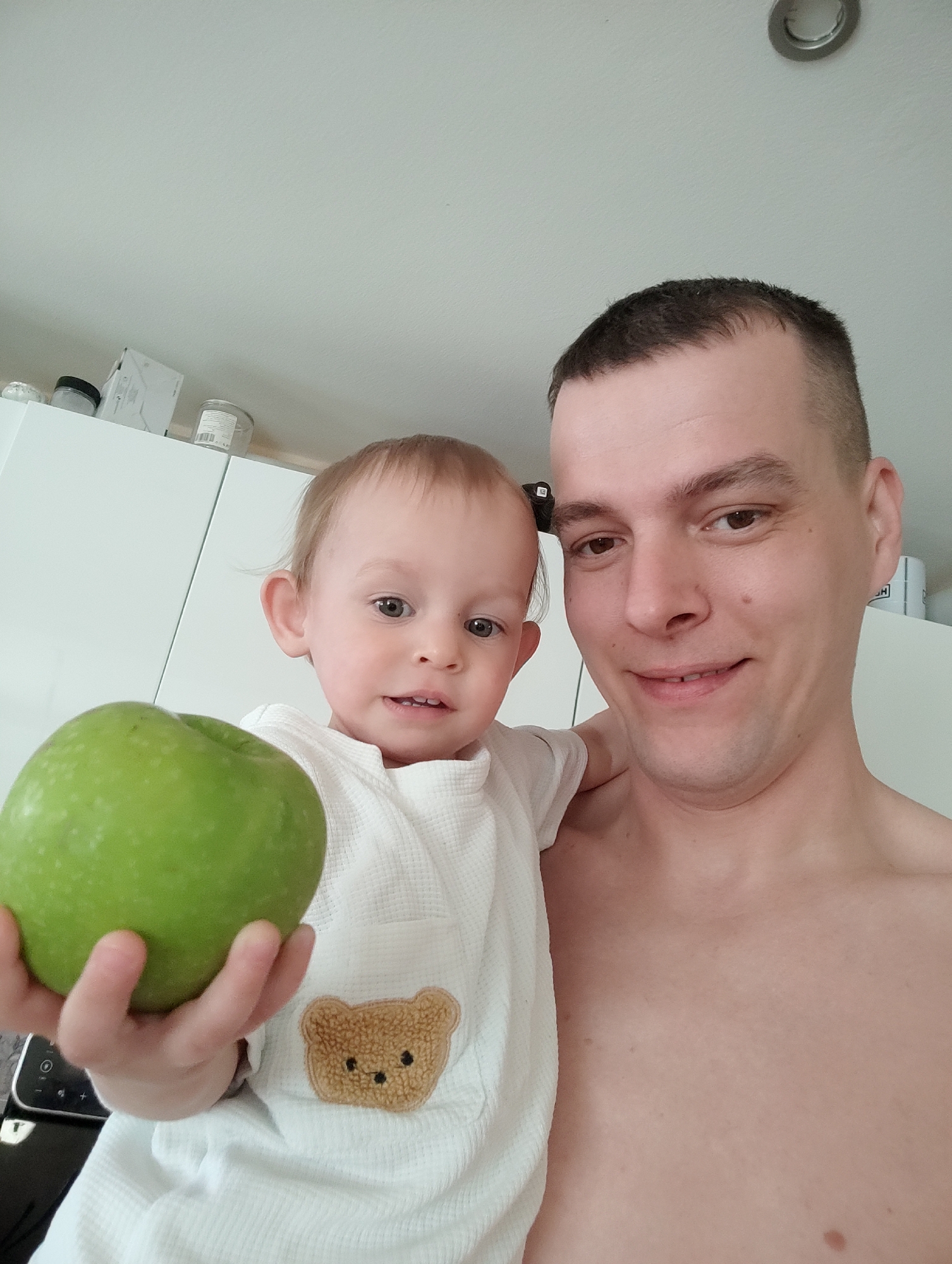 A man and a toddler are posing for a selfie. The man is shirtless and holding the toddler, who is wearing a white robe with a teddy bear applique. The toddler is holding a large green apple in their hand. The background features white cabinets and a ceiling light fixture. The location is in the city of Chaikovsky, Russia.
