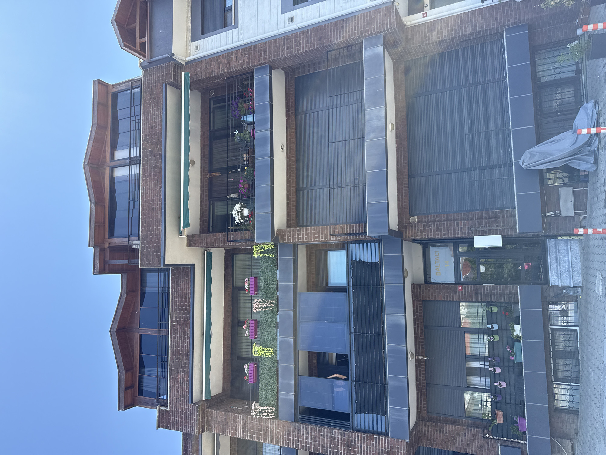 A multi-story brick building with dark shutters and balconies is pictured in Şile, Turkey. The building has several floors with balconies adorned with potted plants and flowers. One balcony on the second floor from the top has a green awning. The top floor features a distinctive wooden roofline with large windows. The ground floor has an entrance with a sign that reads "BALTACI 14". A grey tarp covers an object on the sidewalk in front of the building. The sky is clear and blue.