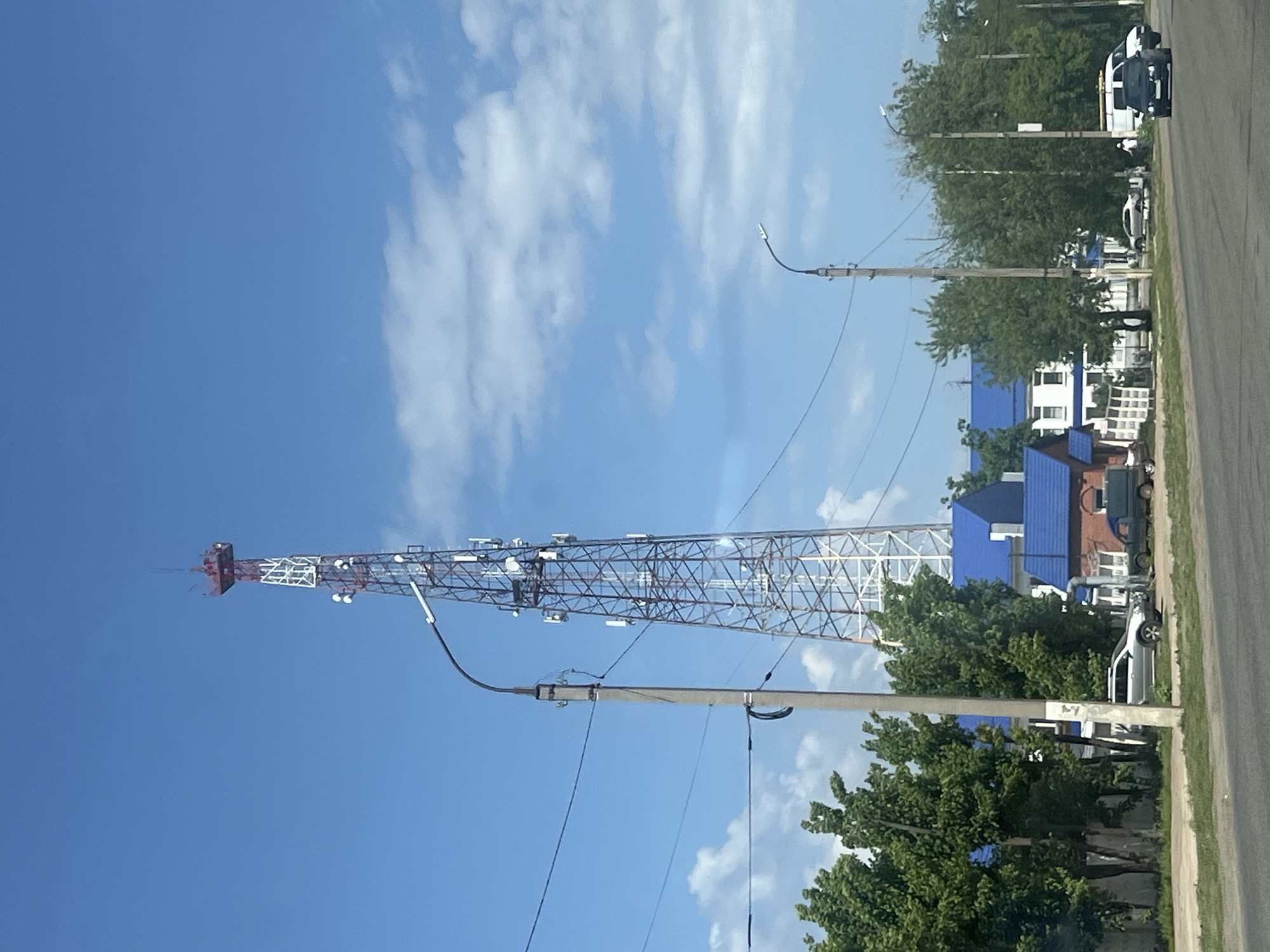 The image shows a cellular communication tower in Chaikovsky, Russia. The tower is red and white, with various antenna arrays attached. Power lines stretch across the lower portion of the frame. A building with a blue roof is partially visible behind trees on the right. A road and several parked cars are present on the right side of the image, suggesting a street scene. The sky is blue with scattered clouds.