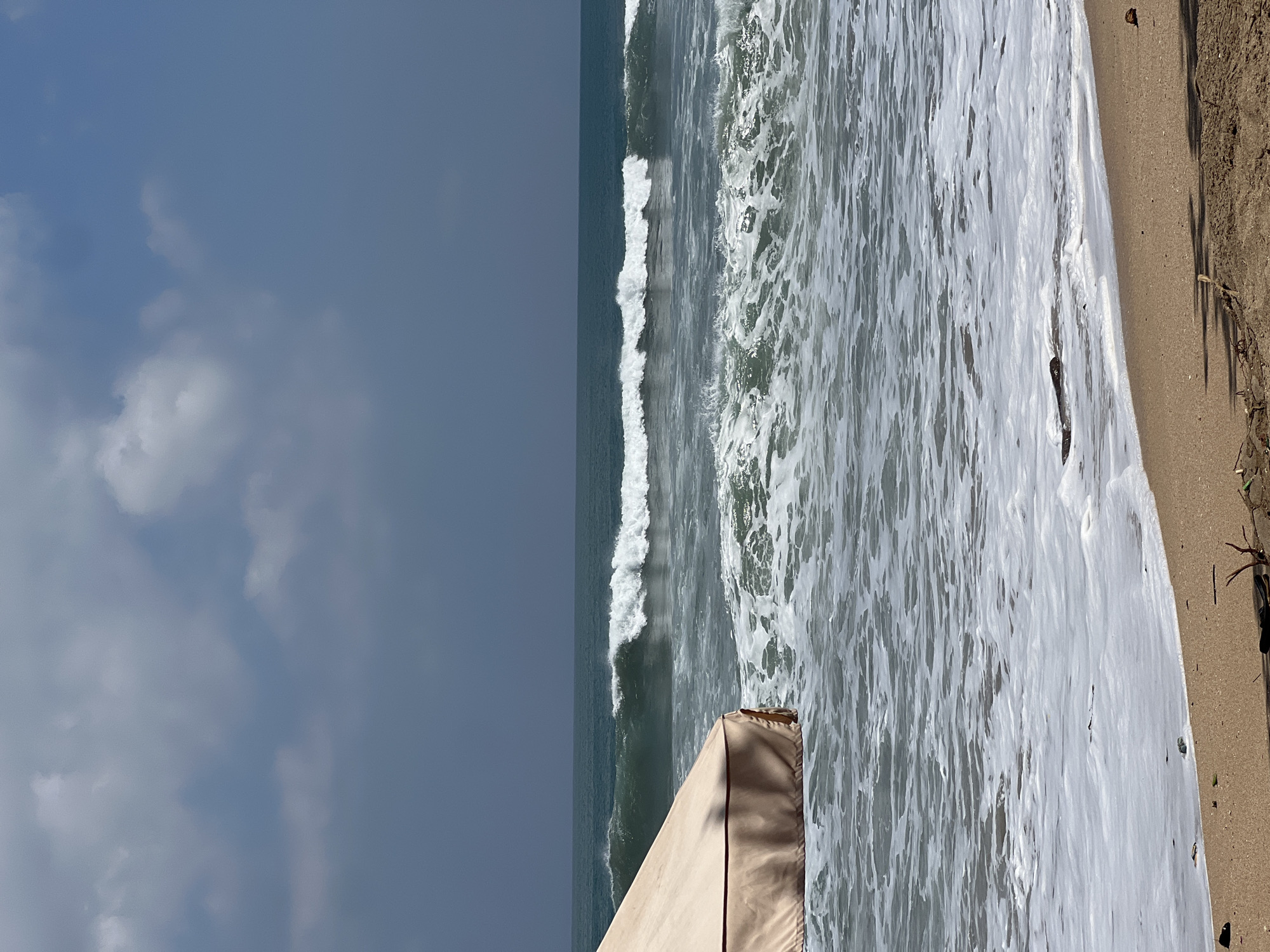 The image depicts a beach scene in Kuta, Indonesia, with a tan-colored umbrella partially visible in the foreground on the left. The ocean is active, with multiple rows of waves rolling towards the shore. The water is a deep teal color with white foam cresting on the waves. The foreground shows wet sand with some debris, including small branches and a piece of driftwood. The sky is a mix of dark gray clouds and lighter blue patches. The overall atmosphere is one of a slightly overcast day at the beach with moderate waves.