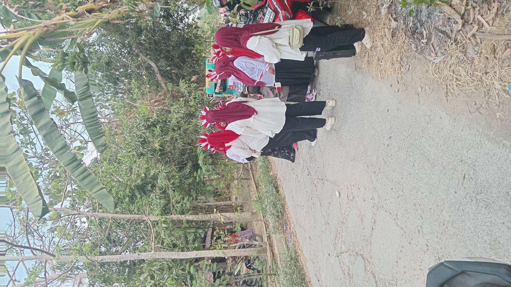 In Kedungwaru, Indonesia, a group of five women dressed in red headscarves and white tops with decorative red and white headpieces stand facing away from the camera on a cracked asphalt road. They are positioned near a red motorcycle and a green cart. The environment is rural, with trees and foliage surrounding the road. One woman in the foreground is wearing black pants and white sneakers, carrying a small beige bag. Another woman behind her is also in black pants and white sneakers. The other women are similarly attired. The scene appears to be outdoors, possibly during a local event or parade.