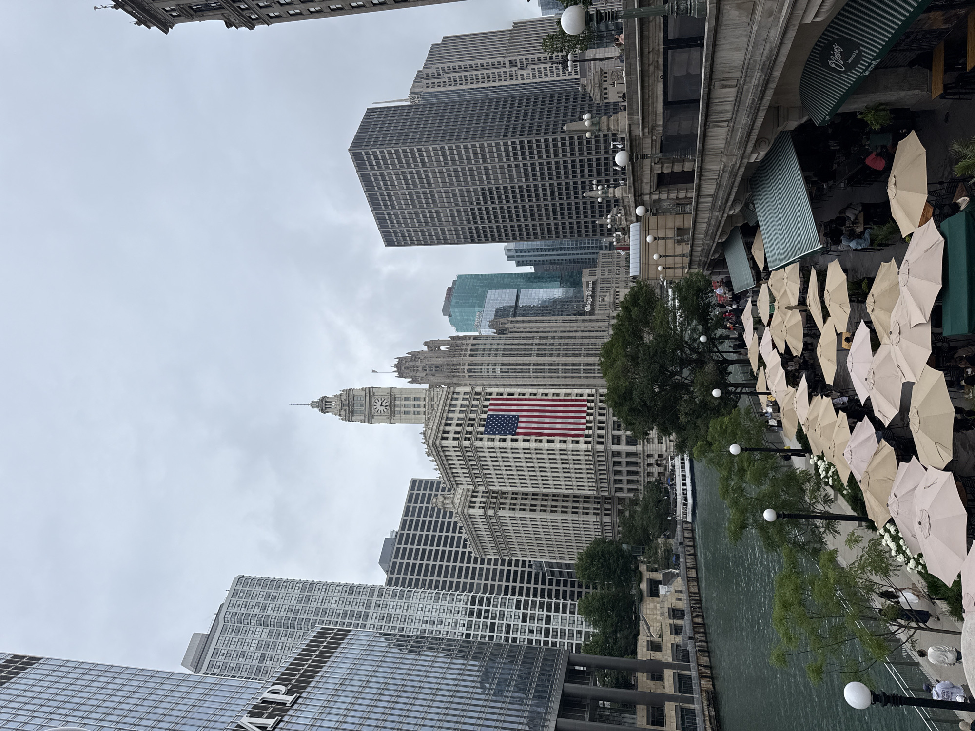 This image depicts an outdoor scene in Chicago, United States of America, likely during daytime under overcast skies. The perspective is from an elevated viewpoint looking down and across a river.
The primary subjects are a collection of buildings, including several tall skyscrapers with distinct architectural styles, ranging from modern glass facades to older, more ornate structures. One prominent building features a clock tower and a large American flag displayed on its facade. Along the riverbank, there is a walkway lined with numerous outdoor tables and umbrellas, suggesting a restaurant or cafe area. Green trees and white flowers add natural elements to the urban landscape. A boat is visible on the river.
Several people are present, appearing as small figures scattered along the riverwalk, some seated at the tables under umbrellas, and others walking. The activity appears to be a casual daytime scene, possibly people dining outdoors or enjoying the riverfront.
The overall mood is subdued, likely due to the overcast weather. The buildings suggest a bustling urban environment, characteristic of a major city. There is some visible text on the awnings of the establishments along the riverfront, though it is difficult to read clearly. The presence of the American flag and the iconic architecture points strongly to a location within Chicago.