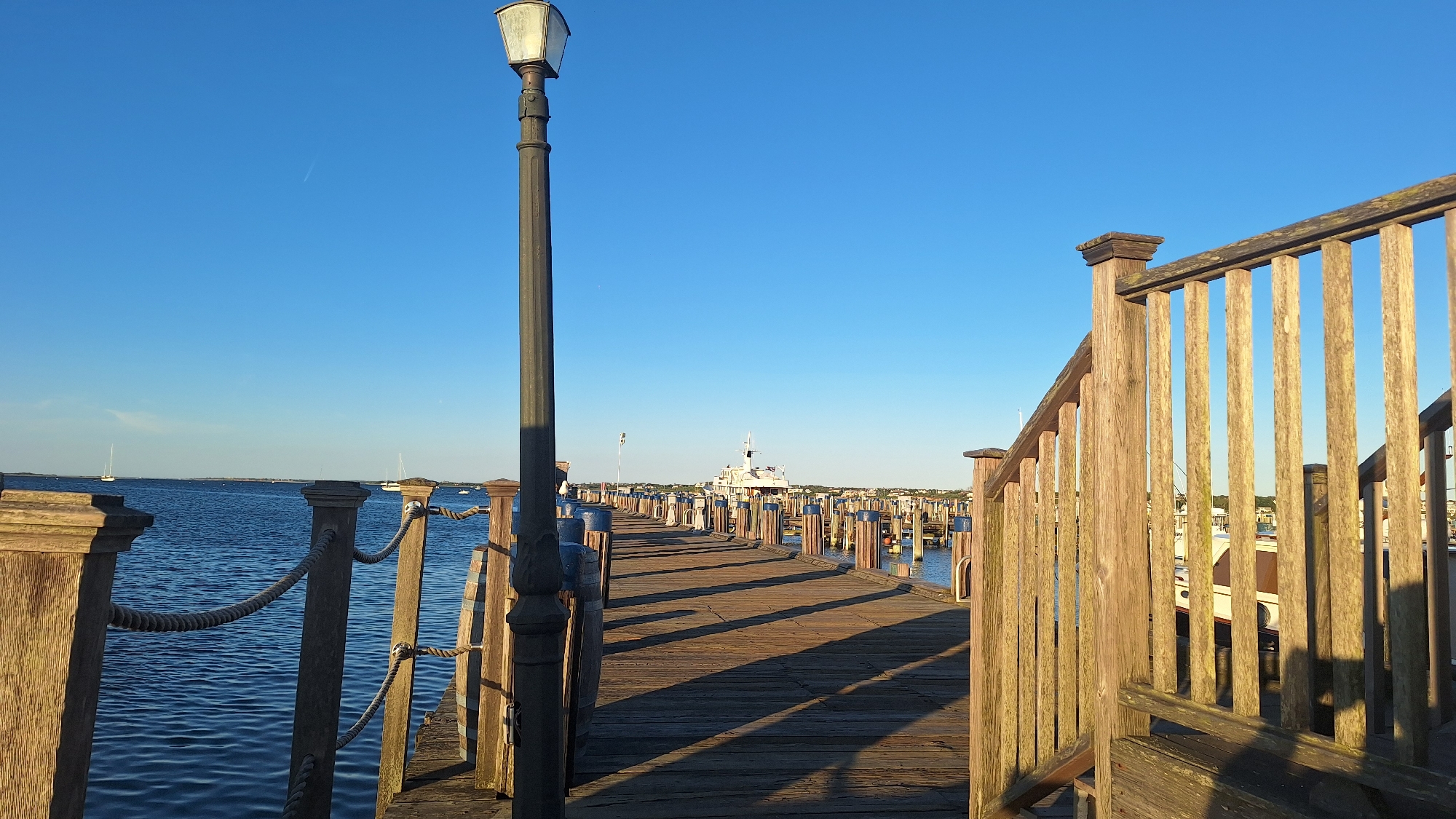 This is a photograph of a wooden dock extending into a body of water. The dock has a wooden railing on the right side and a lamppost on the left side. The water is calm and blue, with a few sailboats visible in the distance. On the far side of the water, there is a shoreline with buildings. The sky is clear and blue, indicating a sunny day. The scene appears to be in Nantucket, United States of America.