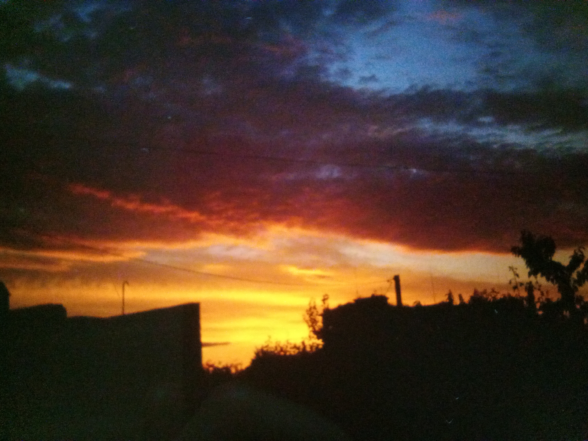 The image shows a silhouetted landscape against a dramatic sky at dusk. The sky transitions from a deep blue at the top to vibrant orange and yellow hues closer to the horizon, with streaks of red and purple clouds scattered throughout. In the foreground, the silhouettes of buildings and trees are visible. A tall, thin pole or antenna stands on the left side of the frame. The overall impression is that of a serene, albeit slightly ominous, sunset over an urban or semi-urban area. The specific location within Myrhorod, Ukraine, cannot be determined from the image alone.