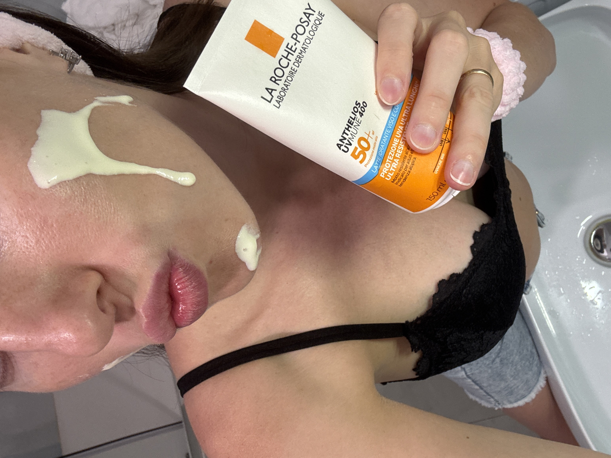 This is a close-up, overhead shot of a person applying sunscreen to their face. The subject is positioned in a bathroom setting, with a white sink visible in the background, suggesting the activity is part of a skincare routine. The person is wearing a black lace bra and denim shorts. Their face is partially visible, with prominent lips and a nose. A dollop of light yellow sunscreen has been applied to the cheek and upper lip area.
The person is holding a tube of "LA ROCHE-POSAY ANTHELIOS UVMUNE 400" sunscreen, indicated by the visible text on the packaging. The tube specifies "50+ SPF" and "PROTEZIONE UVA ULTRA LUNGHI," highlighting its high sun protection factor and broad-spectrum coverage. The text also notes "LATTE IDRATANTE VISO E CORPO" (hydrating face and body milk), "ULTRA RESISTENTE" (very resistant), and that it is "Indicato per adulti e bambini con pelle a tendenza atopica" (indicated for adults and children with atopic-prone skin). The tube contains "150 ml" of product.
Notable details include a silver ring on the person's ring finger and a pink scrunchie on their wrist. The lighting suggests it is daytime, likely indoors. The overall impression is one of personal care and protection from the sun.