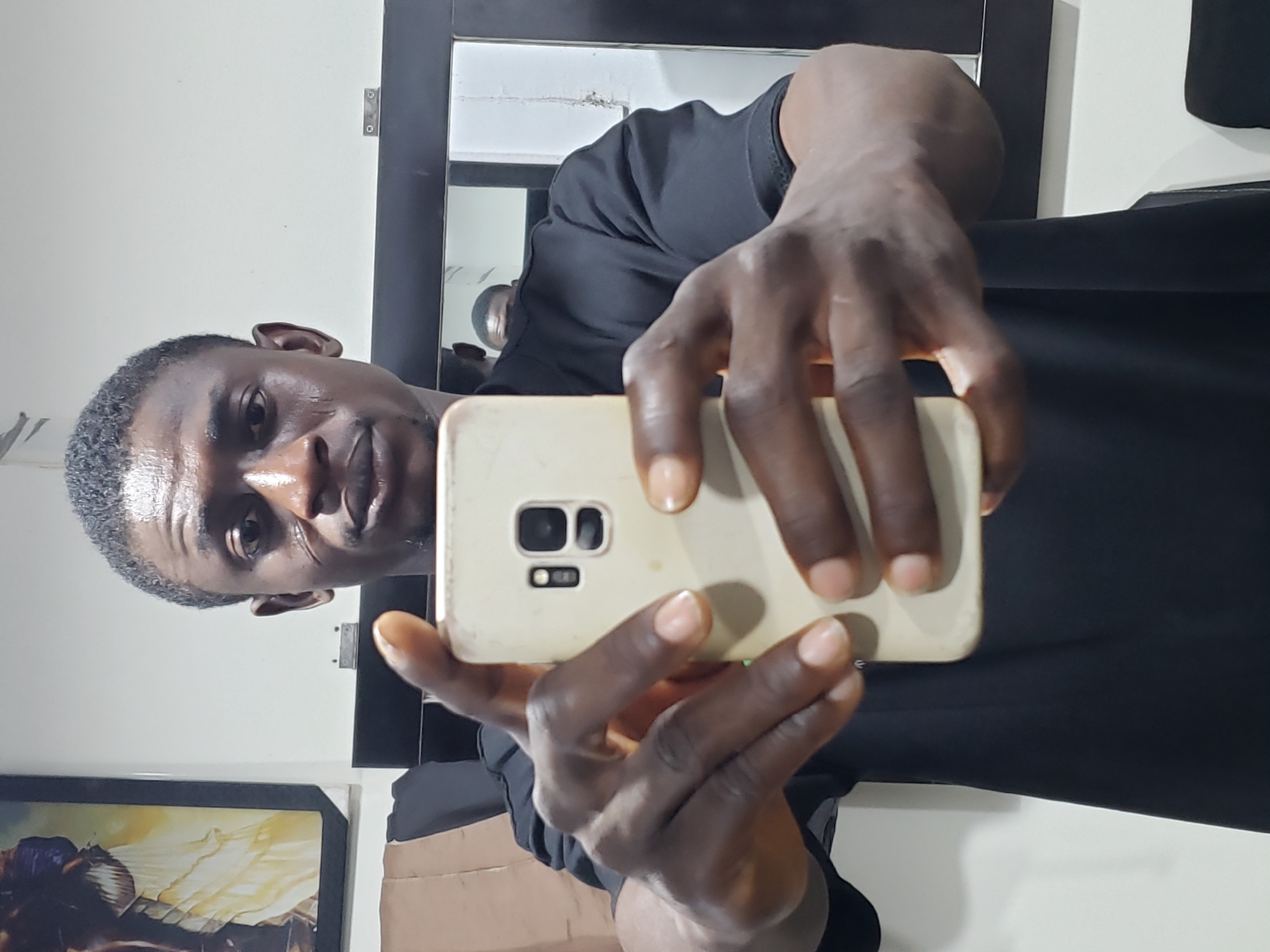 A man is taking a selfie with his mobile phone. He has dark skin, short hair, and is wearing a black t-shirt. The phone appears to be white or light-colored. Behind him, there is a mirror mounted on a wall. The location of this photo is unknown, specifically in relation to the city of Adankolo, Nigeria.