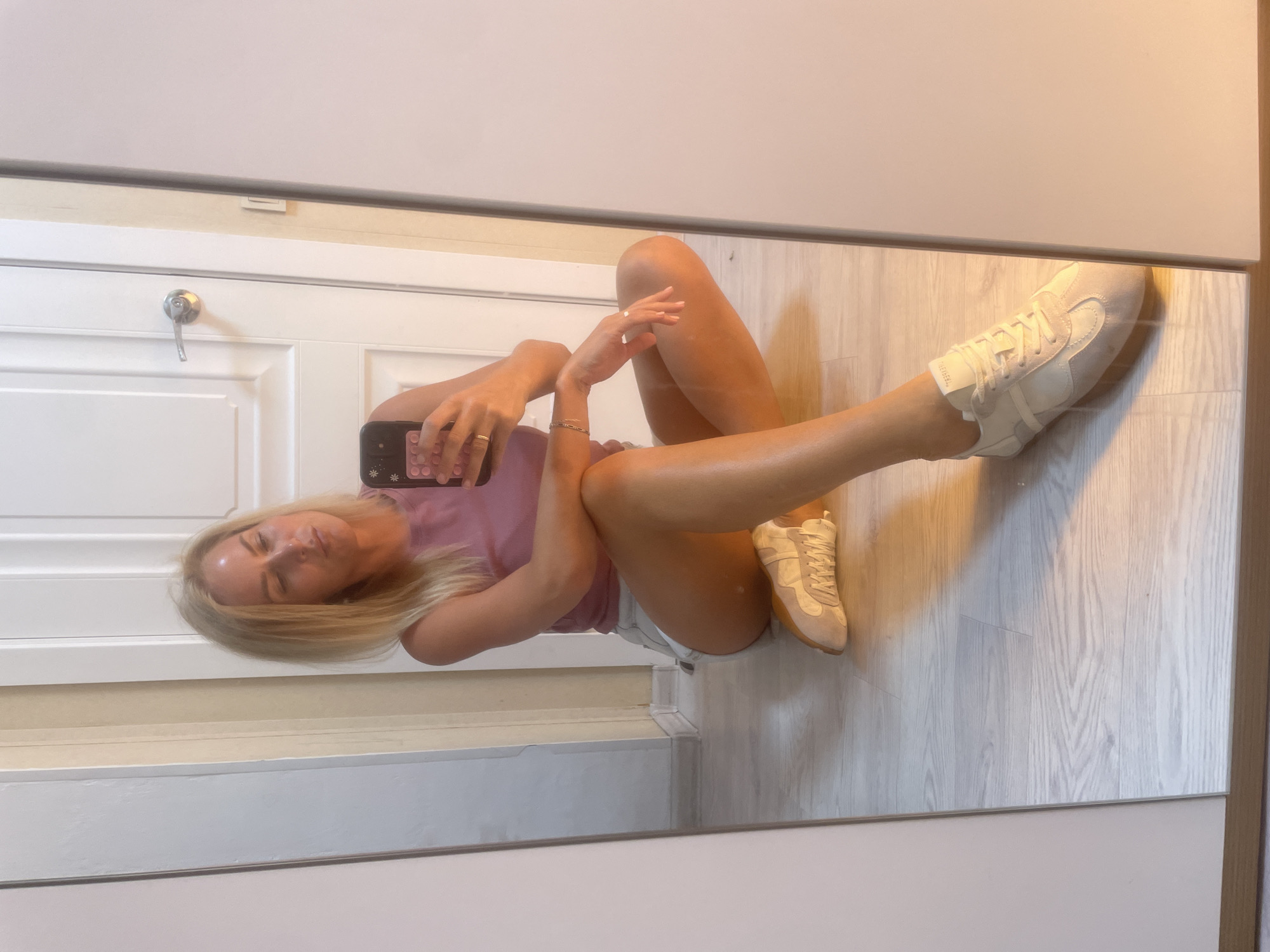 A blonde woman with fair skin and a light complexion sits on a light-colored wooden floor in a room. She is wearing a dusty rose colored tank top and light-colored denim shorts. Her left leg is bent and crossed over her right leg. Her right leg is extended forward, with her foot in a light-colored sneaker. She is holding a smartphone in her right hand, taking a selfie in a full-length mirror. The mirror reflects a white paneled door with a silver doorknob. The background includes a white baseboard and a door frame. The room appears to be indoors, likely a bedroom or dressing area, given the presence of the mirror and door. The lighting is warm, suggesting natural light from a window or artificial lighting. The image does not contain any specific identifiable landmarks or characteristics that would place it in Baranovka, Russia, or any other specific city.