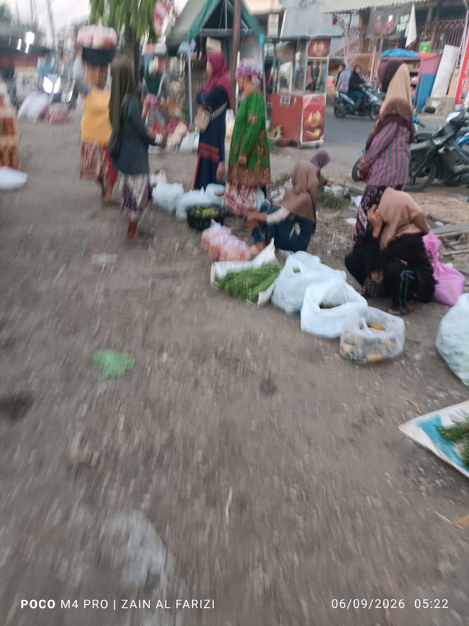 The image depicts a bustling street scene in Arosbaya, Indonesia, likely a market. Several individuals, predominantly women wearing hijabs and traditional attire, are engaged in activities related to commerce. One woman in the foreground is carrying a large basket of goods on her head. Others are seen interacting with each other and with various items laid out on the ground. Scattered across the scene are several plastic bags, some filled with produce, and other market-related items. The ground is unpaved, typical of many outdoor markets. In the background, market stalls and a street with vehicles, including a motorcycle, are visible, suggesting a dynamic and active environment. The overall impression is one of daily life and commerce in a local Indonesian setting.
