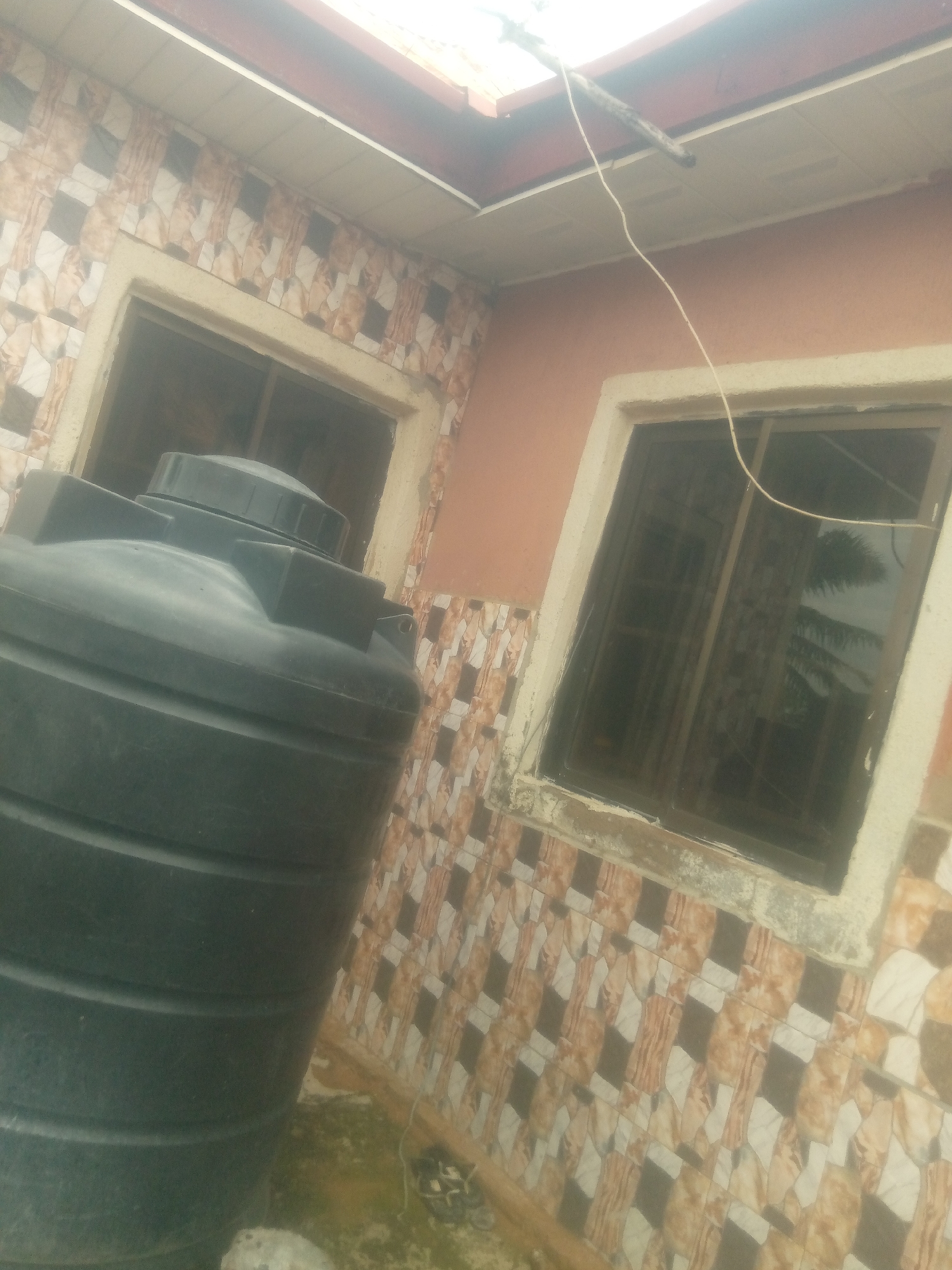 A large, black, plastic water tank is situated on the ground next to the corner of a building. The building has a light orange stucco exterior and is adorned with a patterned tile facade featuring beige, brown, and white geometric designs. Two windows with dark-tinted glass are visible on the building's walls. The window on the left has a beige frame, while the one on the right has a similar frame. A thin, white electrical wire is strung from the building's overhang, passing over the window on the right and extending downwards. The ground surrounding the tank and building is covered in patches of green vegetation and dirt. The image appears to be taken in Jalingo, Nigeria, based on the context provided.