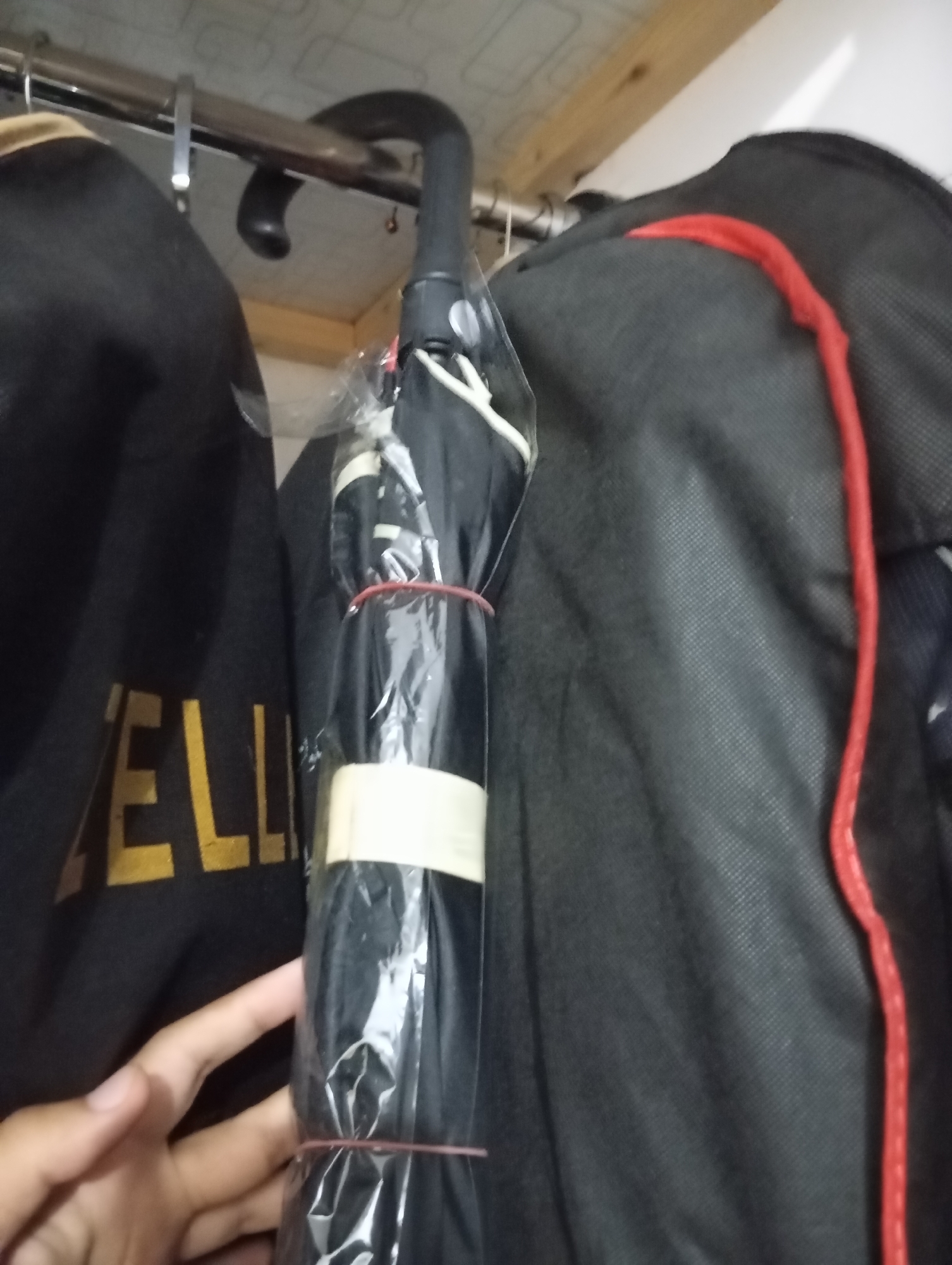 An overhead shot shows a black umbrella with a plastic wrapper and red bands. The umbrella is hanging from a metal rack, and next to it are two black bags with red trim. A hand is visible in the lower left corner of the frame. The scene appears to be indoors, possibly a closet or storage area.