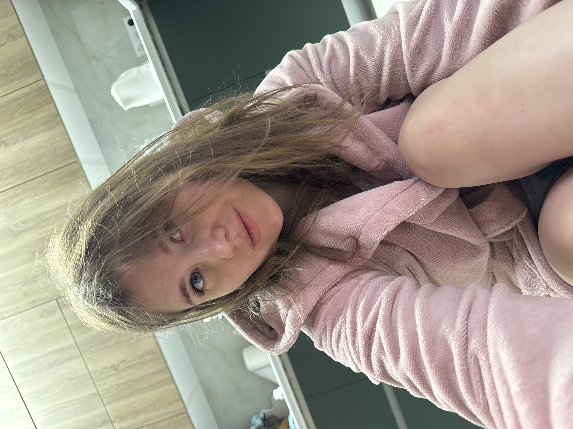 A selfie photograph shows a young woman wearing a pink hooded robe. She is positioned at an angle, looking towards the camera with a neutral expression. Her hair is light brown. The background appears to be a bathroom setting, with tile flooring and a toilet visible in the upper left corner. The location is in Gornyi, Dvurechenskoe S/P, Russia.