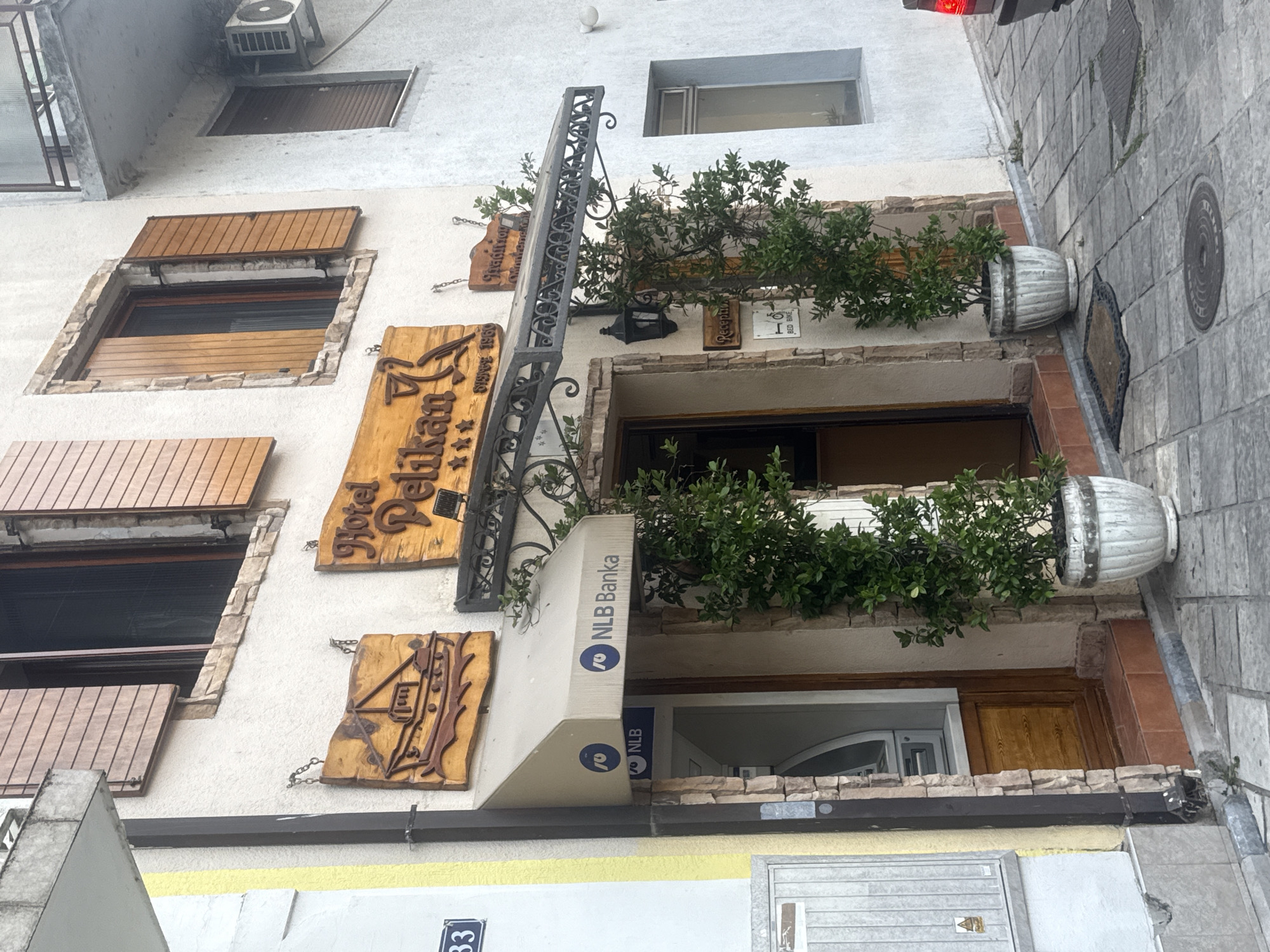 The image captures the facade of a building in Virpazar, Montenegro. A sign for "Hotel Pelikan" with a pelican graphic and three stars is prominently displayed. Below it, a sign reads "Tradition since 1956". To the left of the hotel entrance, an awning with the "NLB Banka" logo and an ATM machine are visible. Two potted green plants flank the entrance. The building features wooden shutters on its windows and is constructed with stone and stucco. The ground is paved with cobblestones, and a lamppost is mounted on the wall. The scene appears to be an outdoor street view of a commercial establishment.