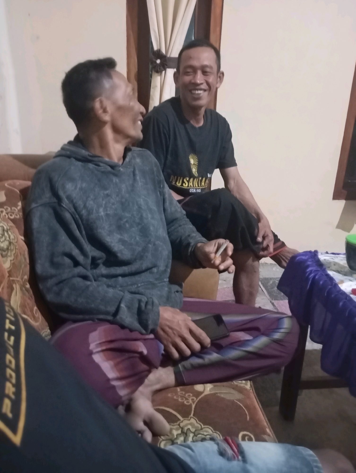 Two men are seated on a couch in what appears to be a domestic setting in Wonogondo, Indonesia. The man on the left, wearing a grey hoodie and a sarong, is holding a cigarette and looking to his left. The man on the right, wearing a black t-shirt with a graphic and dark pants, is smiling and looking towards the camera. A mobile phone is placed on the man on the left's lap. The environment is dimly lit, with a wall and curtains visible in the background.