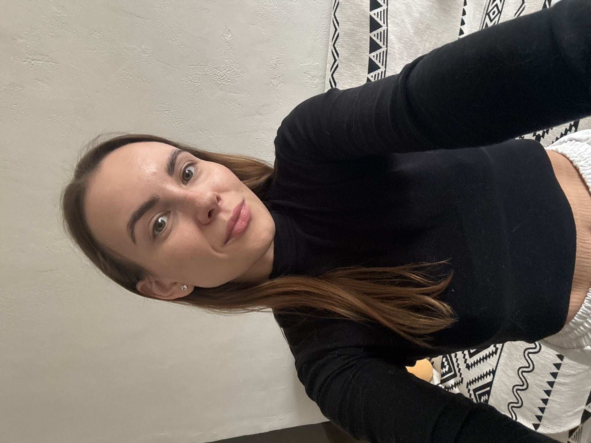 A young woman with brown hair and fair skin is posing for a selfie. She is wearing a black turtleneck sweater and white pants. Her face is visible, and she is looking directly at the camera with a slight smile. The background includes a textured white wall and a white and black patterned fabric. The location is unknown and cannot be determined from the image.
