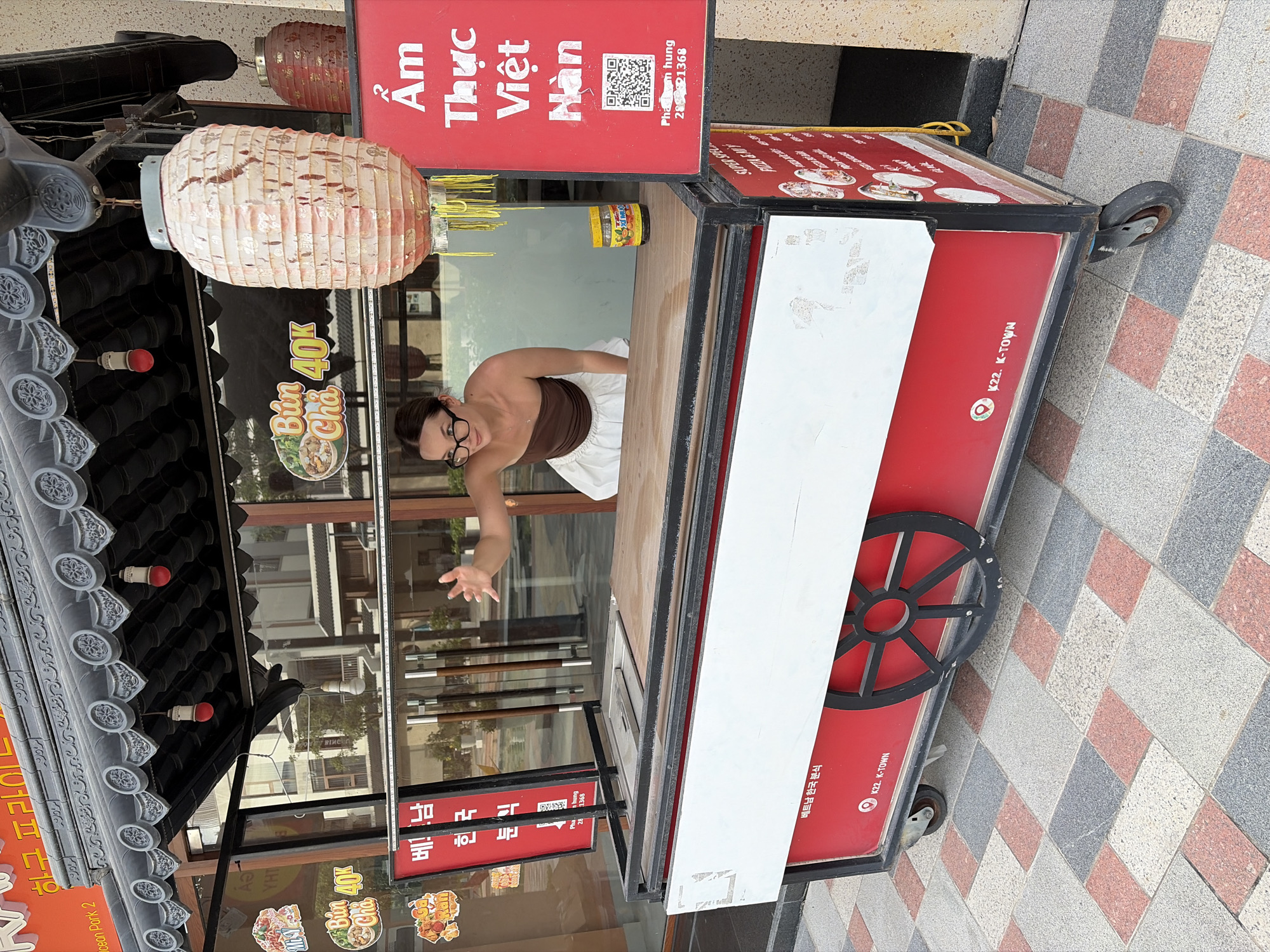 A young woman with glasses and a brown strapless top with a white skirt is standing behind a red and white food cart. She is reaching out her right hand toward the viewer. The food cart has a black metal frame and a wooden countertop. There are various signs on and around the cart, including one with Vietnamese text and a QR code, and another with images of food. A large, slightly damaged paper lantern hangs from the top of the cart. The cart is positioned on a checkered tile surface. In the background, there are reflections of buildings in a glass window.