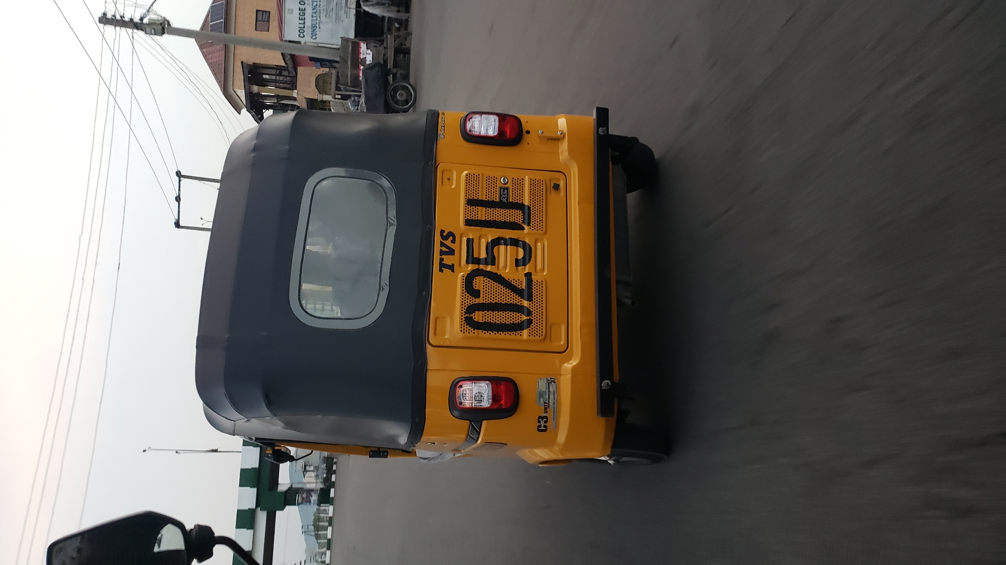 A rear view of a yellow auto-rickshaw is shown as it moves on a paved road, likely in Jalingo, Nigeria. The auto-rickshaw has a black canopy and a prominent license plate with the characters "025JJ". The registration appears to be illuminated by the flash of the camera. The vehicle has a "TVS" logo above the license plate. In the background, a street is visible with some buildings and overhead electrical wires. The sky is overcast. The motion blur indicates the auto-rickshaw is in transit.