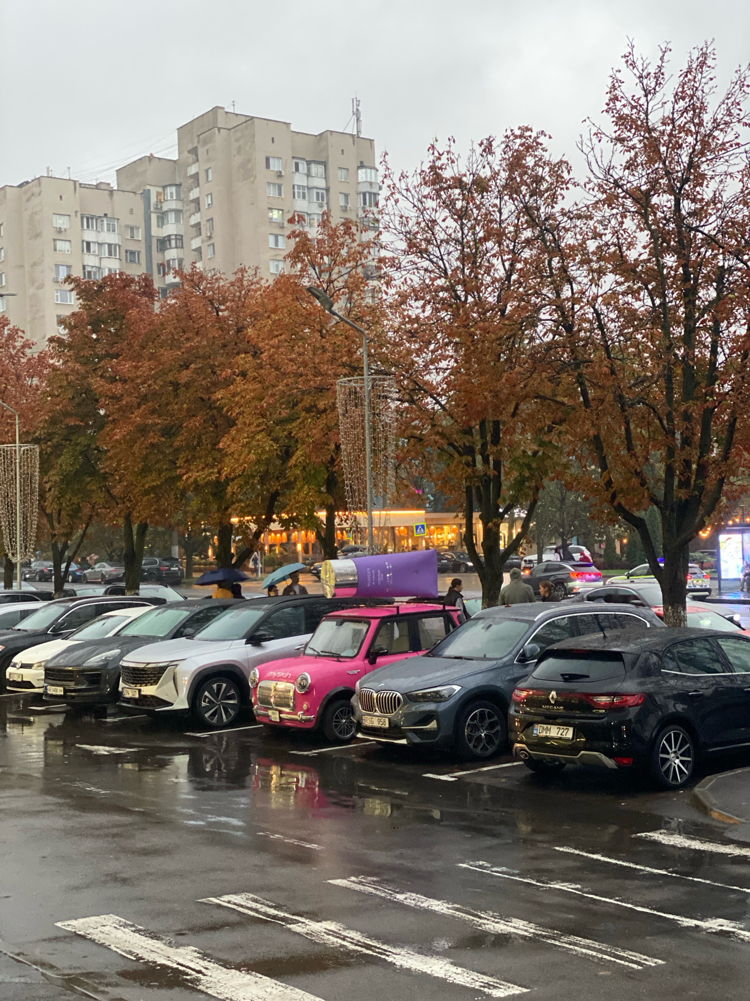 In Chișinău, Moldova, on a wet, overcast autumn day, a street scene unfolds with parked cars and autumnal trees. The foreground is dominated by a row of parked cars, including a pink microcar with a license plate reading "myskin," a dark grey SUV, and a black hatchback with a license plate reading "DMM 727." A pedestrian crossing with white stripes is visible on the wet asphalt. Behind the cars, several trees display vibrant orange and red leaves, characteristic of fall foliage. In the middle ground, more cars are parked, and pedestrians are seen holding umbrellas, one blue and one yellow, suggesting rain. Further in the background, a multi-story apartment building stands under a cloudy sky, with streetlights and decorative lights visible. The overall atmosphere is damp and cool, indicative of a rainy autumn day in the city.