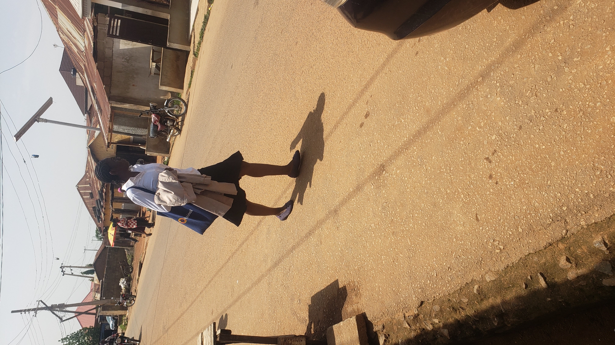 The image depicts a young person walking on a dirt road in Narayi, Nigeria. The subject is wearing a white shirt, a dark skirt, and blue shoes, and is carrying a light-colored bag over their shoulder and a dark blue tote bag. The tote bag has visible text, part of which reads "oil". Behind the subject, several buildings with corrugated metal roofs are visible, suggesting a semi-urban or rural setting. A motorcycle is parked in front of one of the buildings. Further down the road, there are utility poles with visible wires, indicating the presence of electricity. The lighting suggests it is daytime, with a clear sky. The shadow of the person indicates the sun is relatively high, possibly mid-morning or afternoon. The scene appears to be a typical day on a local street.