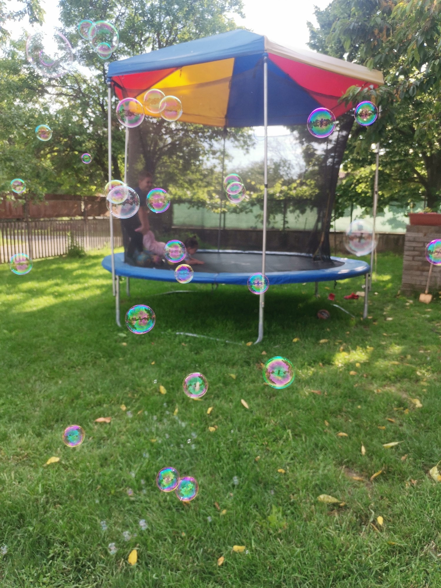 An outdoor scene in Osoitsa, Bulgaria, features a trampoline with a colorful canopy in a grassy yard. Several iridescent soap bubbles are floating in the air, with many concentrated near the trampoline and some scattered across the lawn. A person is visible on the trampoline, and the ground is covered in green grass with scattered yellow leaves. The background includes trees and a wooden fence.