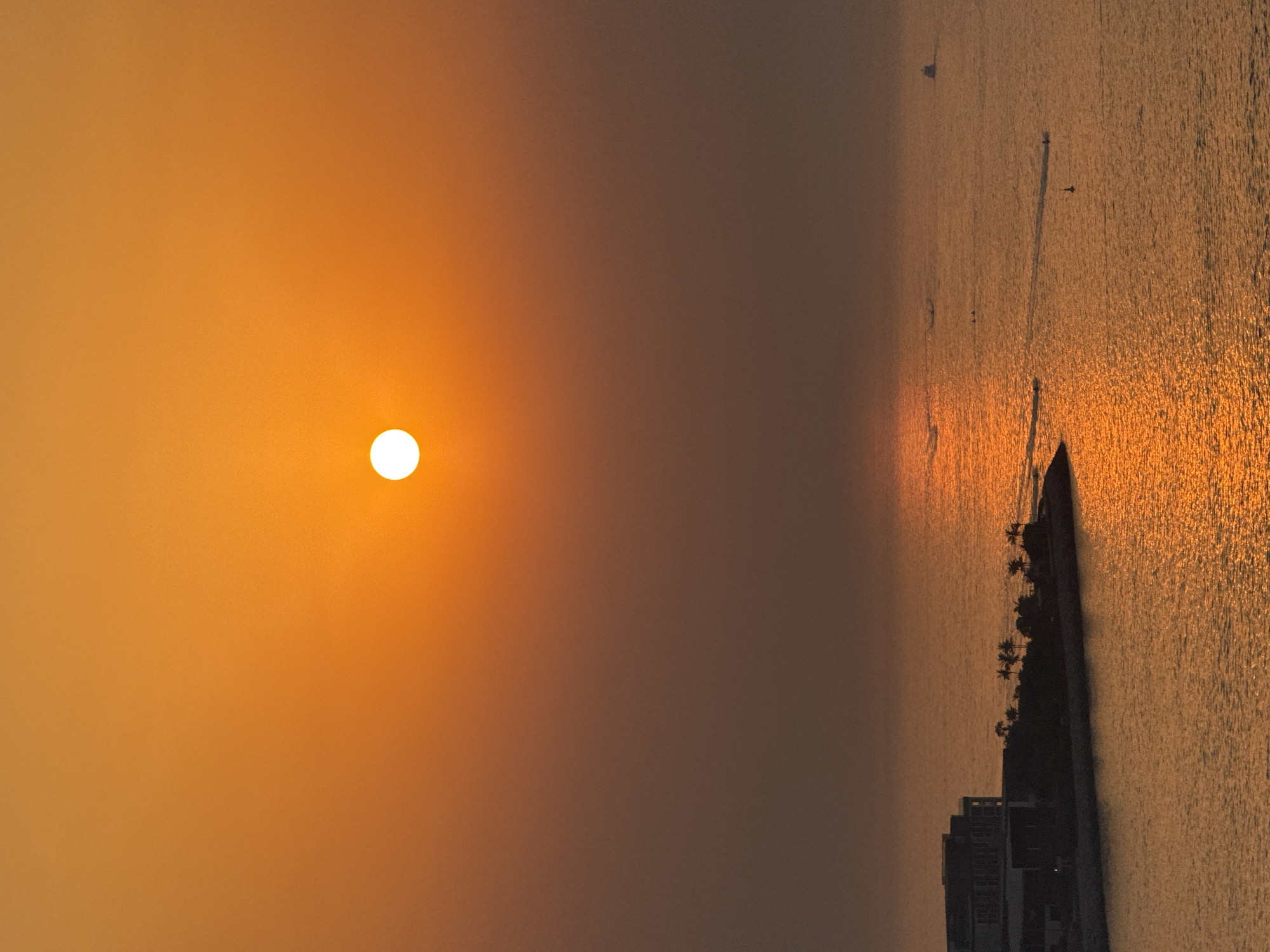 A vertical, high-angle shot captures a sunset over the Arabian Gulf, casting a warm, orange glow across the sky and sea. The sun, a bright, white disc, is positioned in the upper-middle of the frame, its reflection creating a shimmering path on the water. In the lower-left quadrant, a modern architectural structure, likely a hotel or residential building, is silhouetted against the illuminated sea. The building has a multi-tiered, stepped design. Adjacent to it, a strip of land is dotted with palm trees, also silhouetted. Further out in the water, several small boats or personal watercraft can be seen, some leaving trails on the water's surface. The horizon is hazy, obscuring any distant landmasses. The overall scene depicts a serene coastal view in Dubai, United Arab Emirates, during sunset.