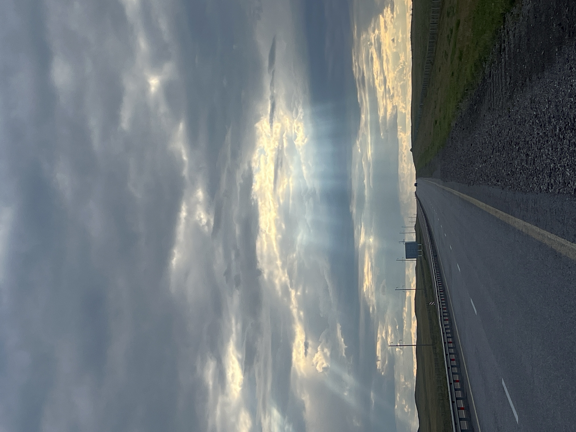 The image shows a straight, paved road in a rural area, likely in the vicinity of Өлеңті ауылдық округі, Kazakhstan. The road has a yellow line marking the edge and white dashed lines dividing lanes. Guardrails run along the left side of the road. The landscape to the right of the road is characterized by rolling green hills, interspersed with grassy fields and a fence line. To the left, more rolling hills are visible, and several utility poles with wires are scattered across the horizon. A large, rectangular blue sign is visible in the distance on the left side of the road. The sky is dramatic and overcast with thick, dark clouds. Shafts of light pierce through the clouds, creating a crepuscular effect over the landscape. A lone vehicle is visible in the distance on the road, heading away from the viewer. The overall impression is one of a vast, open, and slightly somber natural landscape under a dynamic sky.