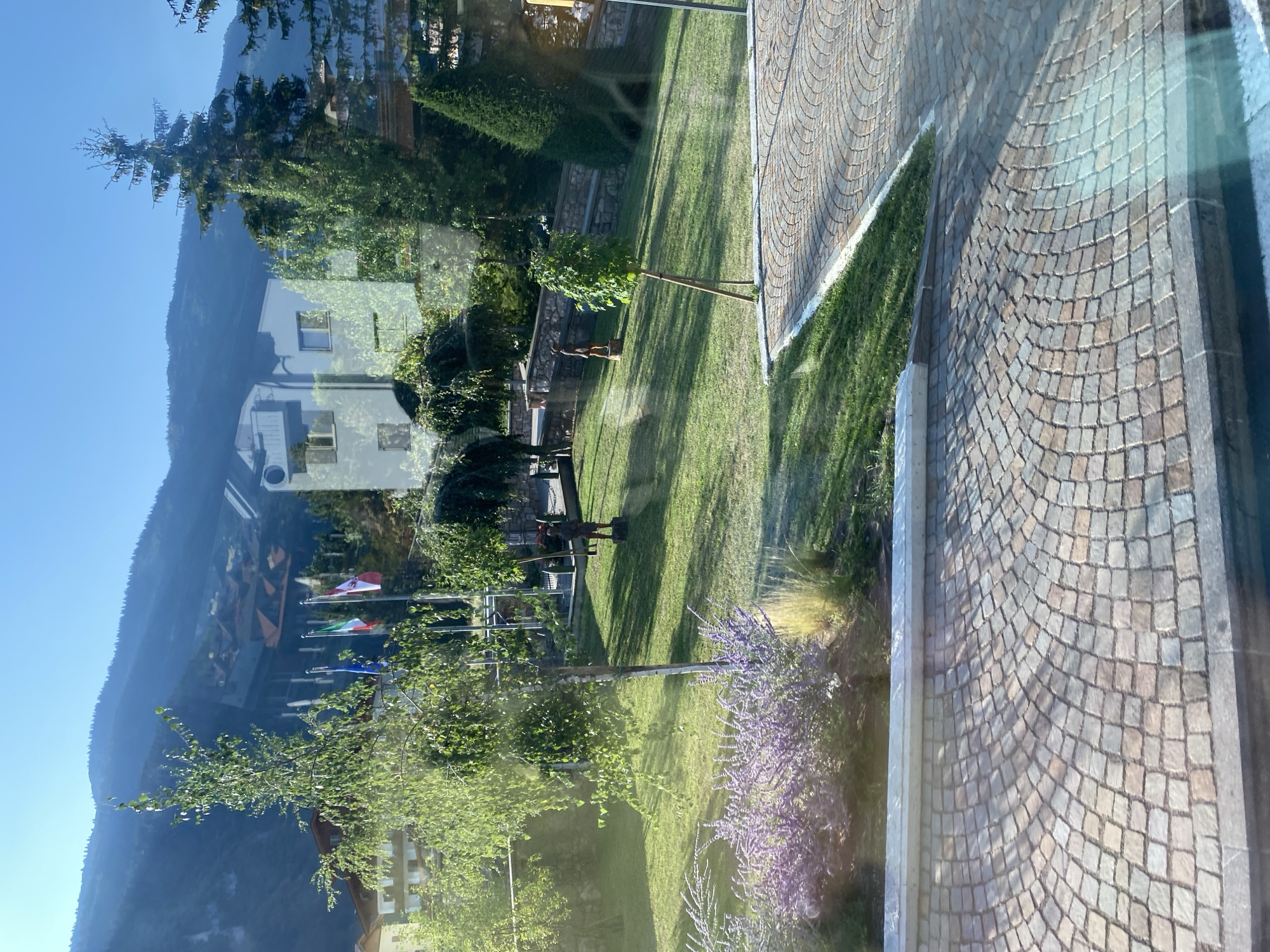 The image captures a scenic view in Molveno, Italy, dominated by lush green lawns, trees, and a backdrop of rolling mountains. In the foreground, a cobblestone path curves into the scene, bordered by a low stone wall and a vibrant patch of purple flowers. Beyond this, a meticulously manicured lawn stretches across the middle ground, dotted with a few young trees and two weathered wooden statues. In the distance, several flags flutter in the breeze, suggesting a public or governmental area. Further back, modern buildings with white facades and dark roofs are nestled against the lower slopes of the mountains, which are covered in dense forest. The sky is a clear, bright blue, indicating a sunny day. The overall impression is one of tranquility and natural beauty, enhanced by the architectural elements that blend harmoniously with the landscape.
