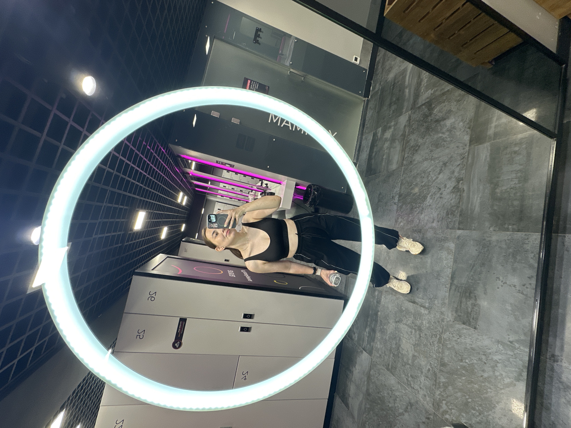 A young woman stands in a locker room and takes a selfie in a large, circular, illuminated mirror. She is wearing a black sports bra and black sweatpants with white stripes. She has a water bottle in her left hand and is holding her phone in her right hand to take the picture. The locker room has white lockers with grey doors. There are also pink lights visible in the background. The floor is tiled with grey, mottled tiles. The ceiling is black and has a grid pattern with recessed lights. The reflection in the mirror also shows a wooden bench or storage unit to the right. The image is taken from a slightly high angle, looking down at the woman's legs and feet.