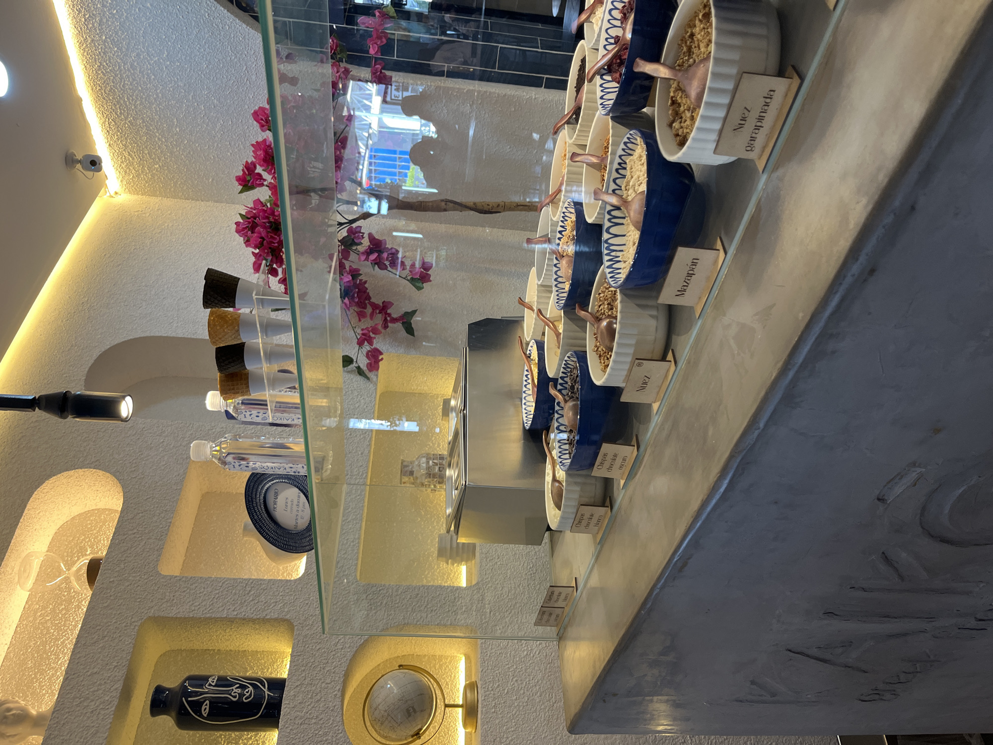 The image, taken in Puebla, Mexico, depicts the interior of what appears to be a dessert or ice cream shop, characterized by a clean, modern aesthetic. The scene is brightly illuminated by warm strip lighting tracing a curved architectural feature on a textured white ceiling and by lights within recessed wall niches. A small white security camera is visible on the ceiling.
In the foreground, a dark grey counter, possibly with embossed text, extends towards a glass display case. Inside the case, numerous white and blue ceramic bowls contain various dessert toppings, each accompanied by a small wooden spoon. Visible wooden labels identify these toppings in Spanish, including "Mazapán" (marzipan), "Nuez garapinada" (candied walnut), "Nuez" (walnut), "Chispas chocolate oscuro" (dark chocolate chips), and "Chispas chocolate blanco" (white chocolate chips). Stacked black and white ice cream cones are also part of the display.
Behind the main display, glass shelves hold other items. One shelf features small bottles of "KAIGO" water and a circular sign displaying the "HORARIO" (Hours): "Lunes cerrado Martes a domin 12-8pm" (Monday closed, Tuesday to Sunday 12-8pm). Below this, a decorative wall features several illuminated, arch-shaped niches containing various objects, including a blue vase with a white abstract face, a small globe, and other sculptural elements. Pink artificial flowers, resembling bougainvillea, add a touch of color to the background. The overall setting suggests a pleasant and inviting retail space.