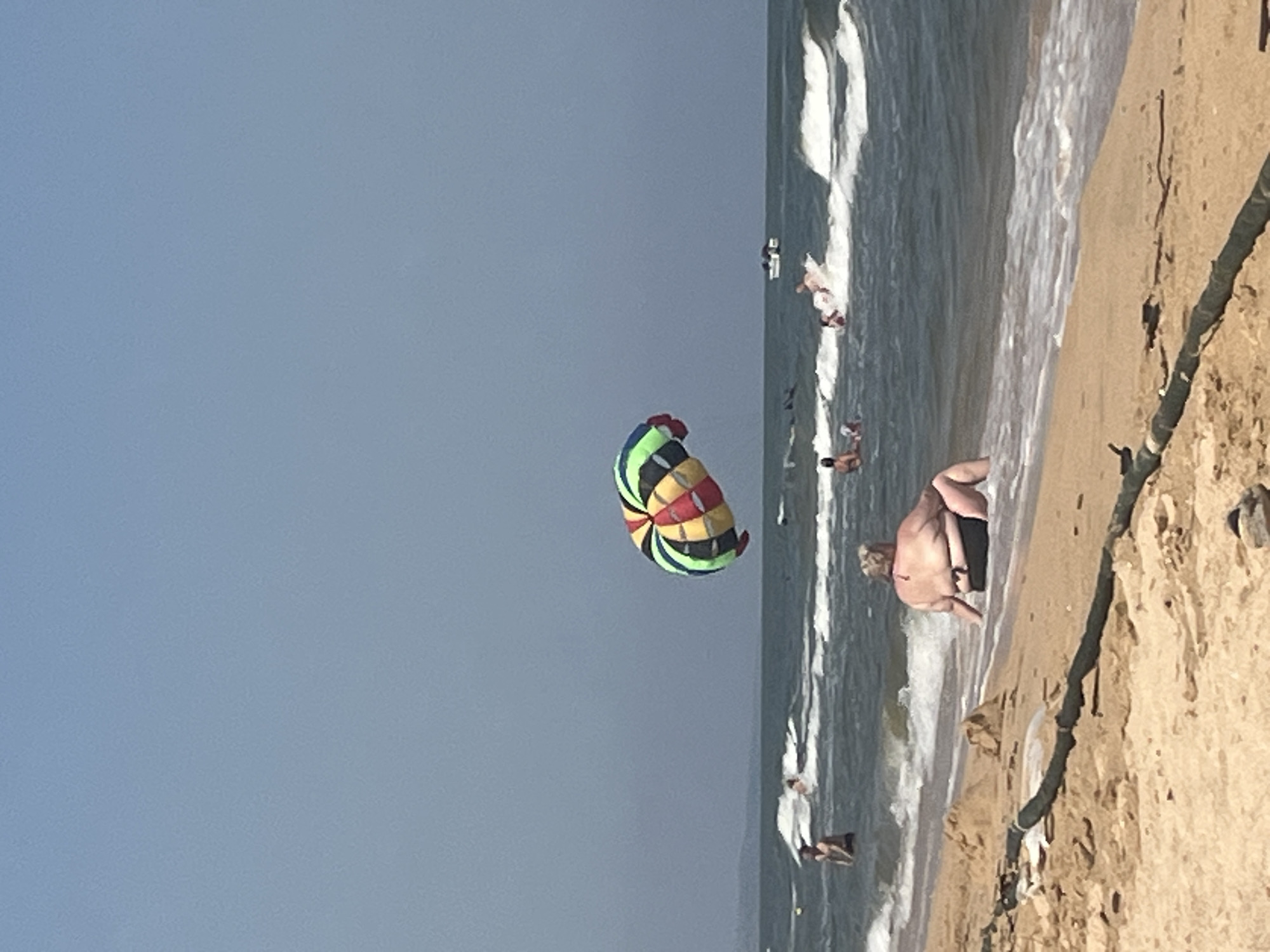 The image depicts a sunny day at a beach in Golubitskaya, Russia. The foreground shows a sandy beach with some debris, including a thick, dark hose-like object. In the shallow water, a woman is sitting, facing away from the camera, wearing a black swimsuit. Beyond her, the sea is active with waves, and several people are visible in the water. To the left, a man stands in the water. Further out, a group of people are enjoying a watersport, possibly parasailing, with a colorful parachute visible in the sky. A boat is also present in the distance. The sky is clear and blue.