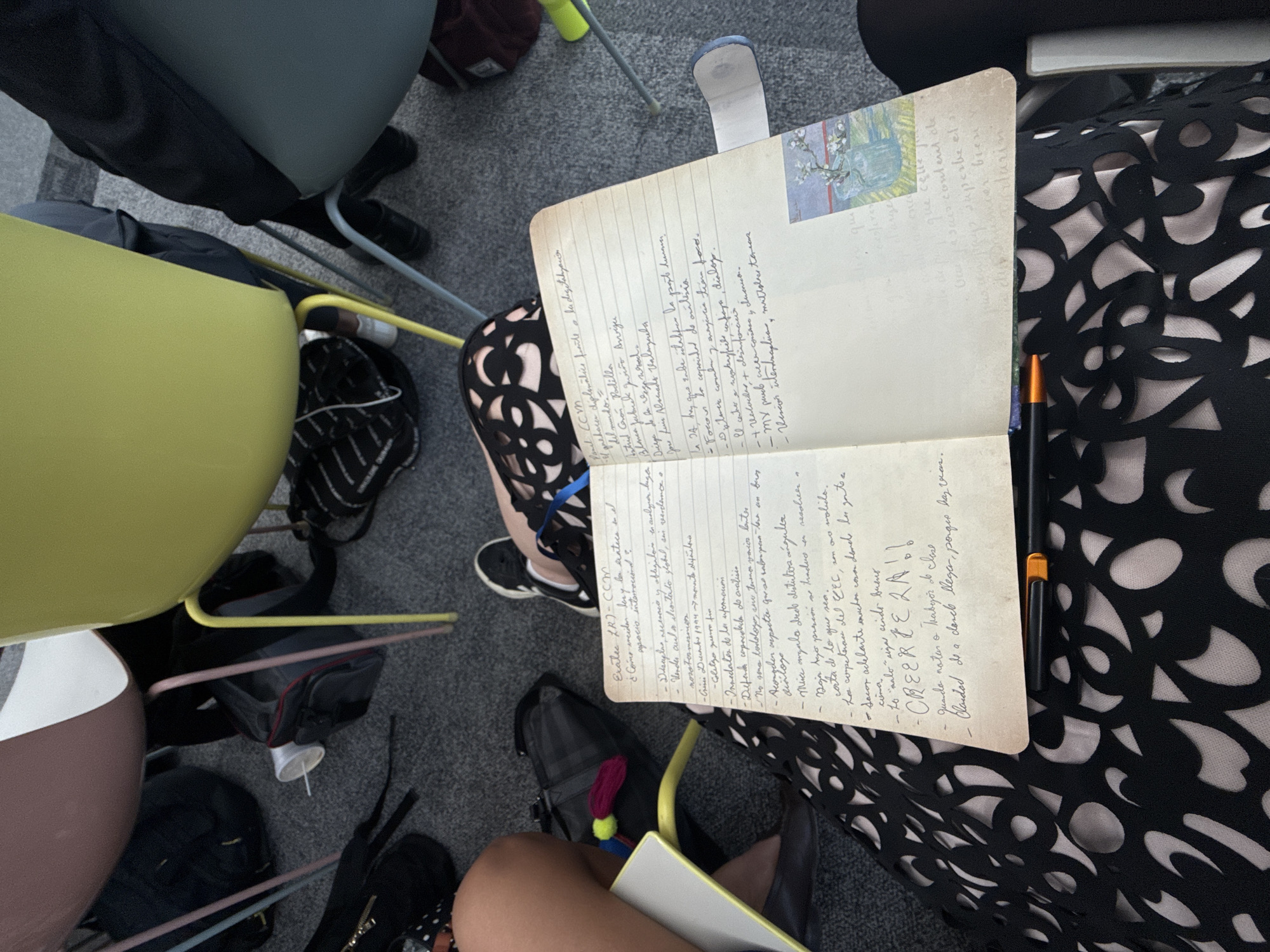 An overhead photograph captures an indoor scene, likely a classroom or lecture environment. The central focus is an open, lined notebook with handwritten text in Spanish, resting on the lap of an individual in the foreground. This person wears a black garment with a complex cut-out pattern over a light-colored layer and black flat shoes; their left leg and thigh are prominent. A black pen with an orange band lies across the open notebook. A small color print, depicting flowering branches in a vase, is affixed to the lower right page of the notebook. To the right, another person's right leg and foot are visible, clad in black pants/leggings and black-and-white sneakers. The floor is covered with grey carpet. Numerous bags, including black backpacks and a grey bag with red trim, are scattered on the floor. The legs of multiple chairs are visible, exhibiting various colors such as pink, yellow, and grey, supporting seats in shades of mauve, light green, and grey. A yellow folder is partially visible near the foreground person's left leg. The image provides no specific visual indicators to identify the location as Mexico City, Mexico.
