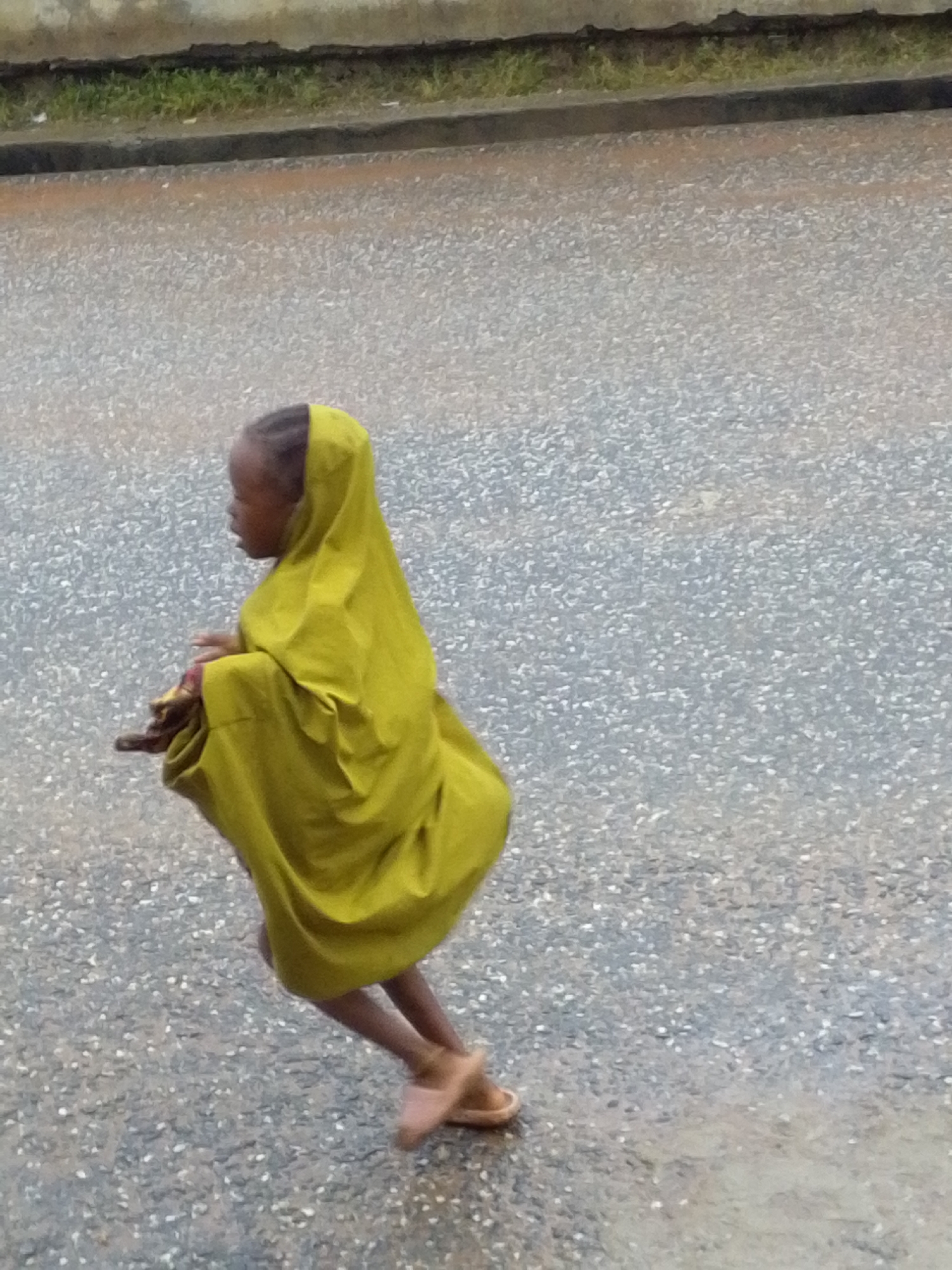 In Serti, Nigeria, a young child walks on a wet, asphalt road. The child is wearing a bright yellow headscarf and a matching garment that drapes over their body. Their face is partially visible, turned to the left. The child is walking with their legs bent, suggesting a hurried pace or a playful stride. Faint shadows are visible on the road, indicating overcast conditions or perhaps the presence of nearby structures casting shade. The background shows a concrete barrier or curb with a strip of green grass beyond it, and a textured wall or building facade. The asphalt is speckled with small, reflective particles, indicating a damp surface.