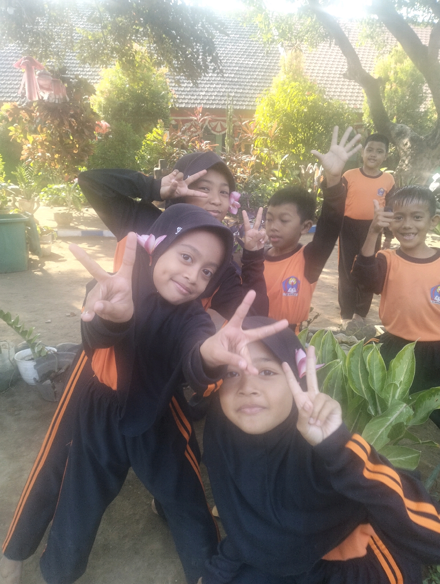 In Blabakan, Indonesia, a group of five children, four girls and one boy, are posing for a photo outdoors. Two girls in the foreground are wearing black hijabs and black tracksuits with orange stripes. The girl on the left is holding up two fingers in a peace sign, and the girl on the right is also making a peace sign. Behind them, a boy in an orange t-shirt and black pants is giving a thumbs-up gesture. To his left, another boy in an orange t-shirt is waving. Further back, a fifth child, also in an orange t-shirt, is raising their hand. The background features lush green plants, trees, and a building with a tiled roof. The lighting suggests it is daytime, possibly late morning or early afternoon, with the sun casting a bright glow. The ground appears to be dirt or sand.