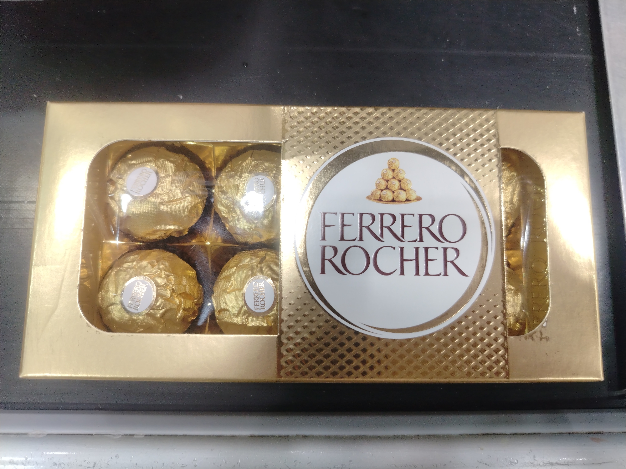 A box of Ferrero Rocher chocolates is displayed on a dark, possibly granite countertop. The box is gold with a clear plastic window revealing four chocolates. The chocolates are individually wrapped in gold foil, each with a white circular label displaying "FERRERO ROCHER". A circular emblem on the box features a pyramid of these chocolates and the brand name "FERRERO ROCHER" in dark red lettering. The background is out of focus, but appears to be a kitchen or dining area. The lighting is bright and even, suggesting indoor illumination. There are no people or any other objects of note visible in the image. The scene is static and posed, likely for product display or marketing purposes.