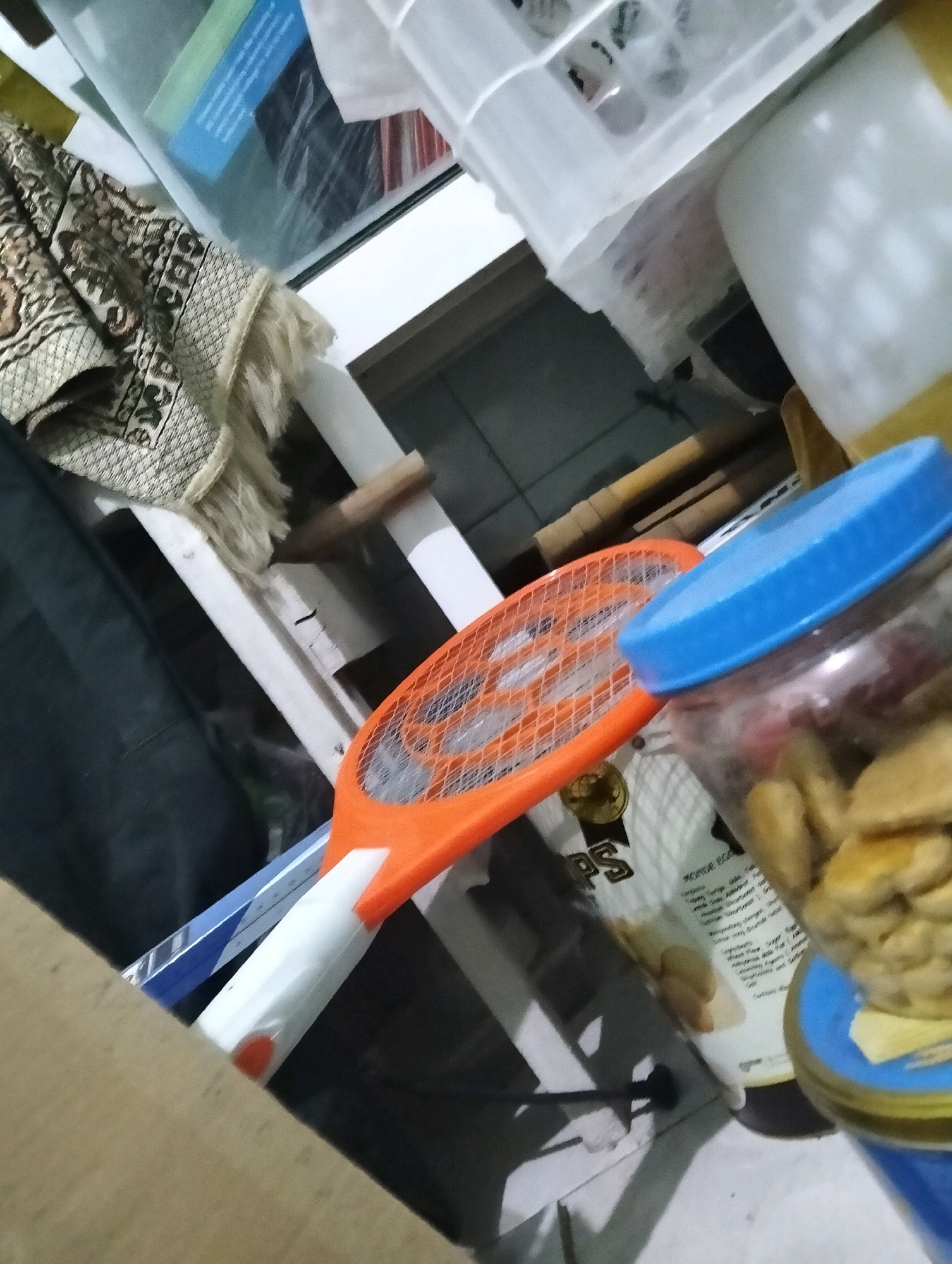 The image depicts a close-up, cluttered indoor scene. In the foreground, a transparent jar with a blue lid is filled with light-brown, irregular-shaped objects, possibly snacks or dried fruits. To the left of the jar is an orange and white electric mosquito swatter, its mesh head facing upwards. Behind these items, a white shelf unit is visible, with various containers and objects stacked on it. A patterned textile, possibly a rug or tapestry, is draped over the left side of the shelf. The background shows more shelving and various household items, suggesting a storage area or a room with limited space. The lighting is dim, with some areas brightly illuminated, creating shadows. The overall impression is of a disorganized but functional domestic space. The scene is not identifiable as being in Paciran, Indonesia, or any specific city.